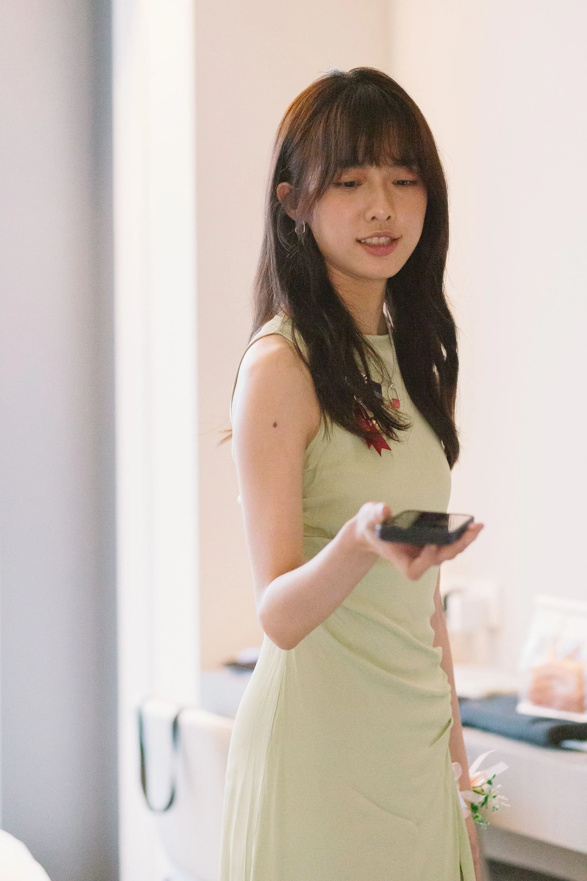A young woman with dark hair, wearing a light-colored dress, holding a smartphone, standing indoors, with a blurred background.