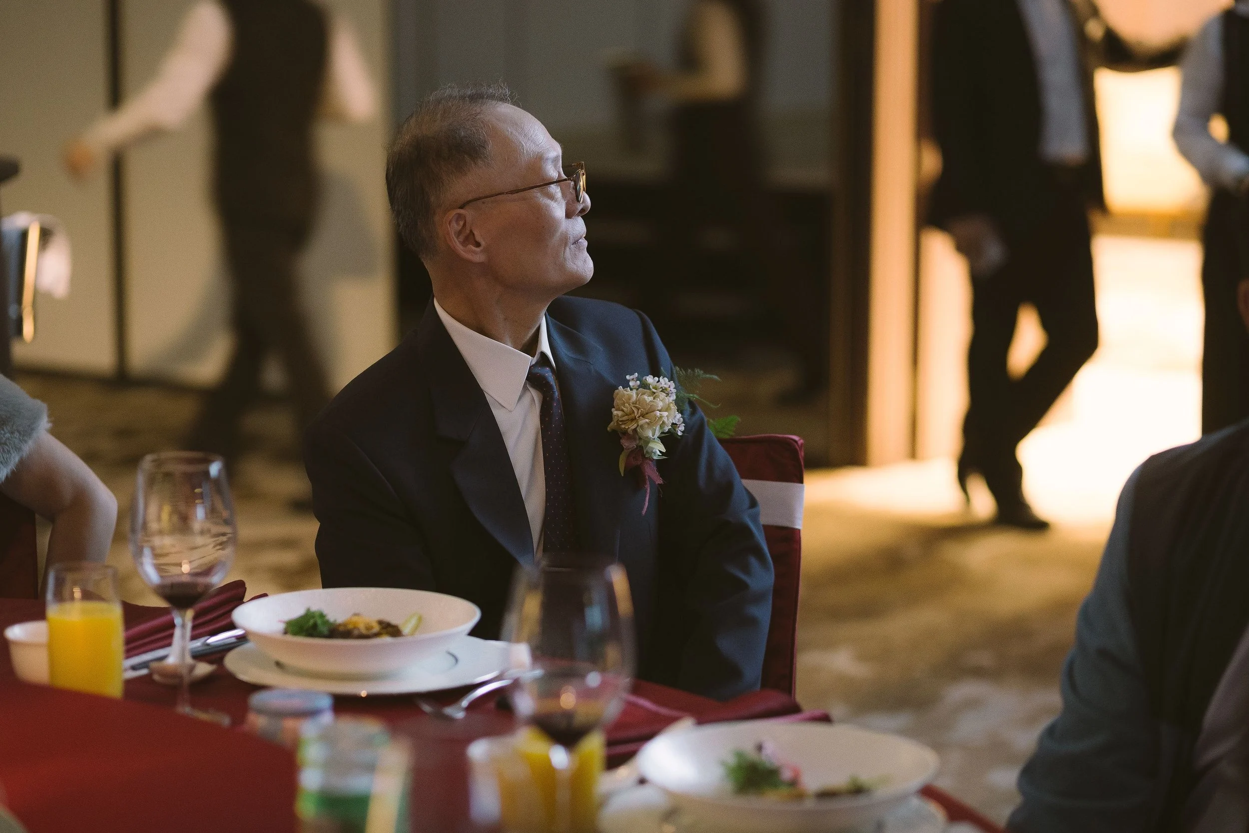 A man dressed in a suit with a boutonniere sitting at a dining table during a formal event or celebration.