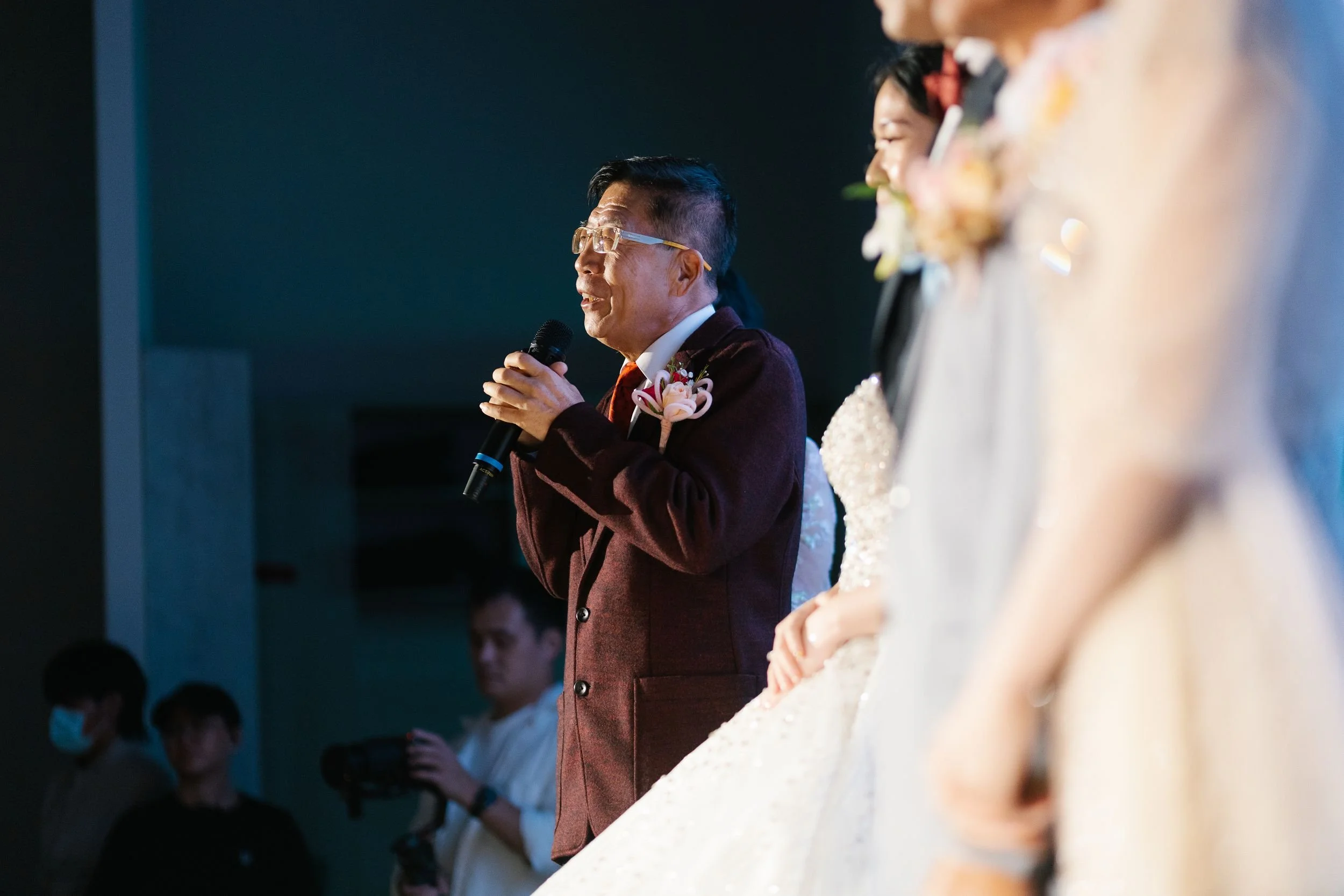 A man in glasses and a maroon suit jacket speaking into a microphone at a formal event, with people in formal attire, including a woman in a white dress, visible next to him.