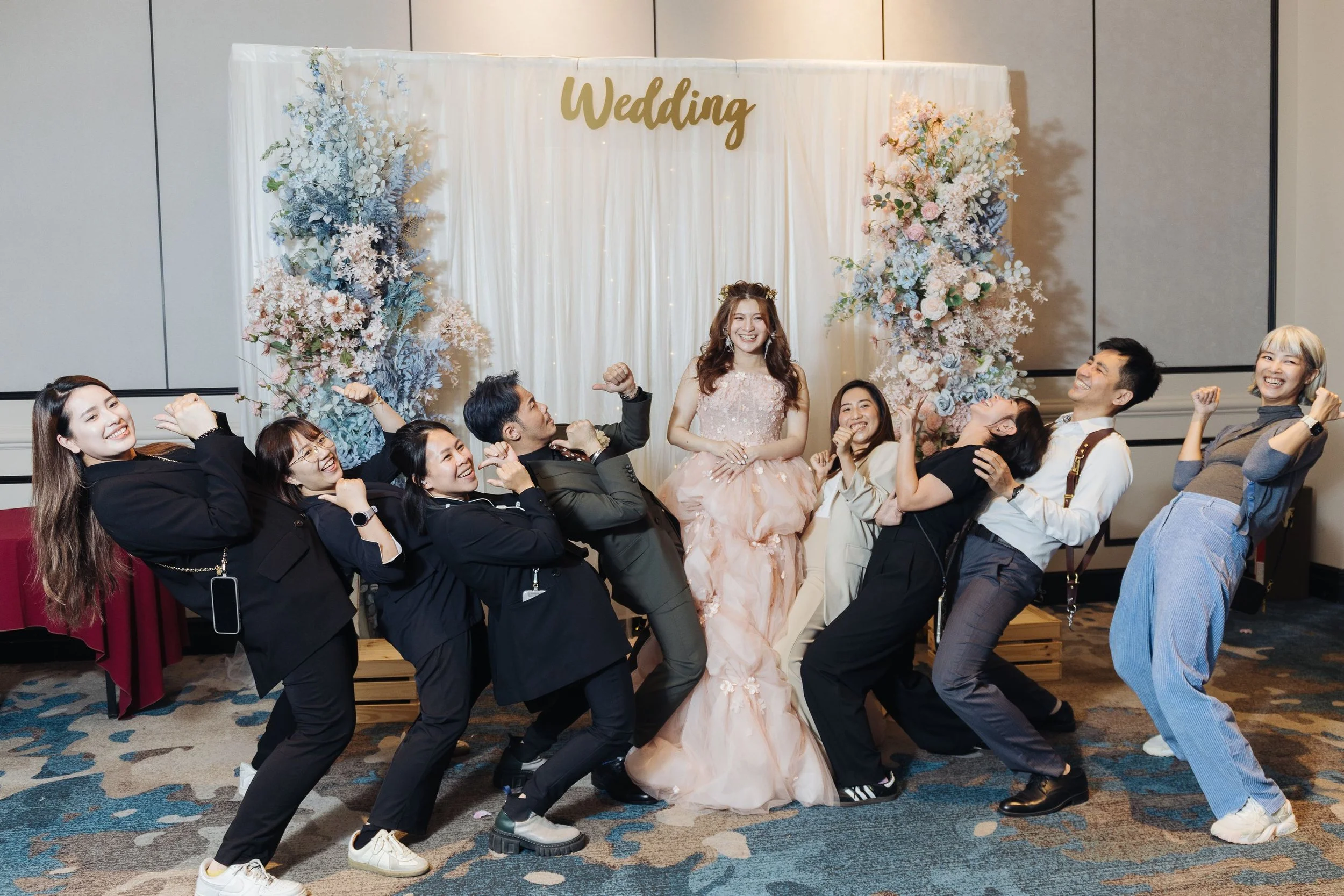A group of people celebrating at a wedding reception, posing playfully in front of a decorated backdrop with the word "Wedding" in gold letters, surrounded by large floral arrangements.