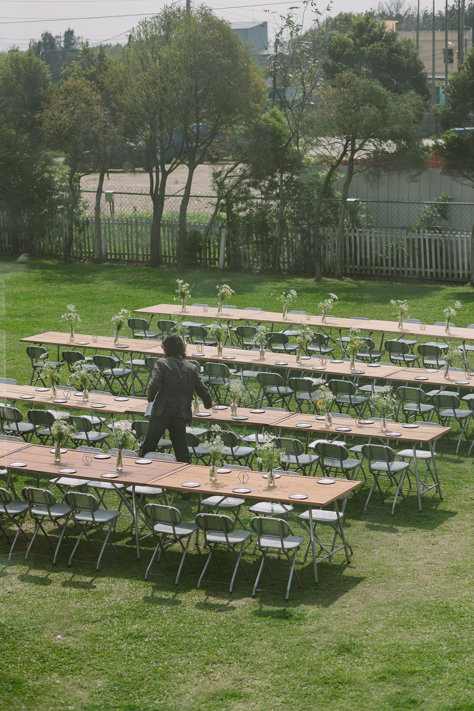 Empty outdoor banquet setting with long tables, chairs, and small vases of flowers on a lawn, with trees, a fence, and parked cars in the background.