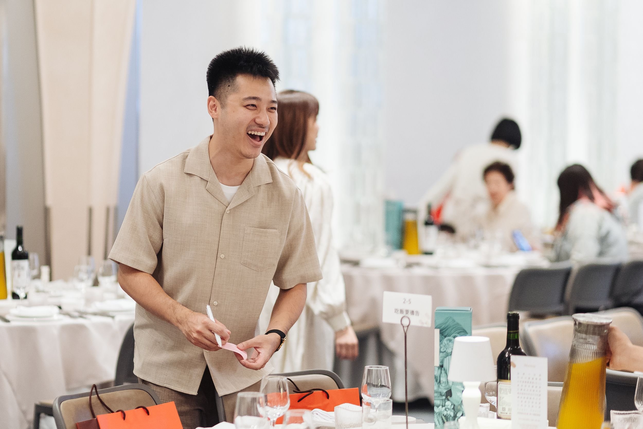 A young man with short black hair smiling while holding a pen in a banquet hall decorated for a celebration or event; other people are in the background.