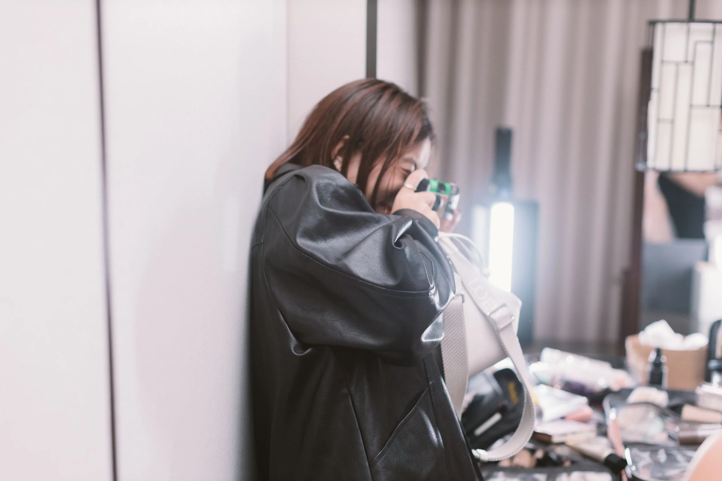 A woman with brown hair leaning against a wall, holding a smartphone close to her face, smiling.