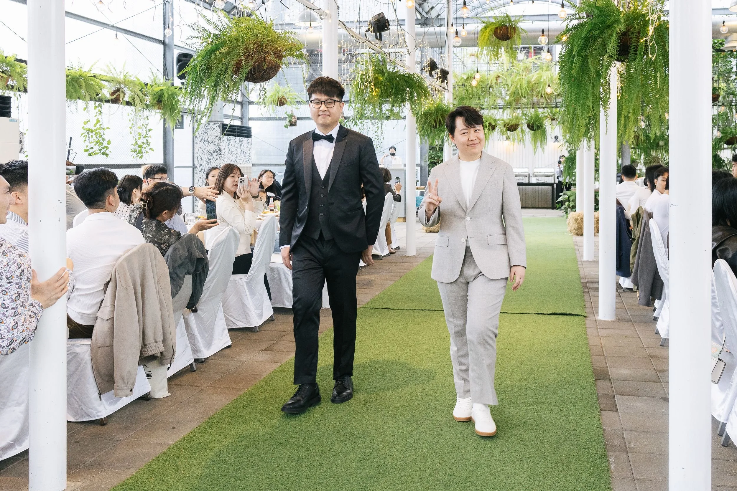 Two people walking down an aisle at a formal event or wedding, with one person in a light gray suit making a peace sign and the other in a black suit with a bow tie. Guests seated on either side of the aisle are taking photos or watching. The setting
