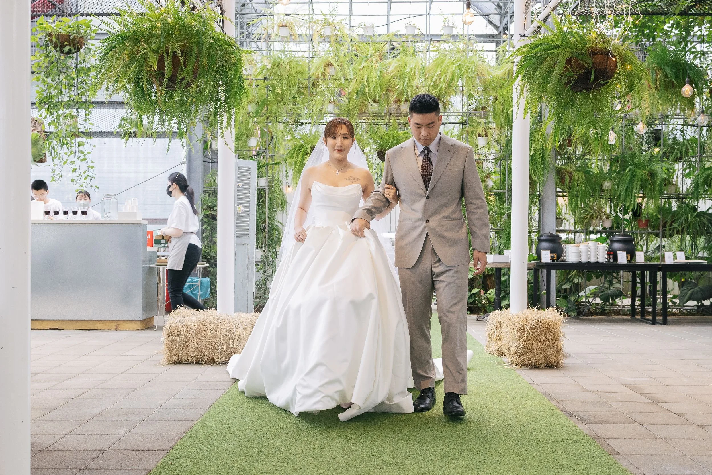 A bride in a white wedding dress and veil walking with a groom in a light grey suit inside a greenhouse decorated with hanging plants and string lights. Two women in white shirts and black masks are in the background near a counter, and hay bales are