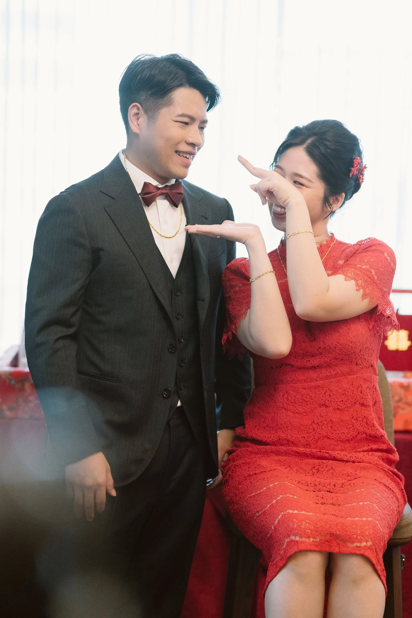 A man in a tuxedo and a woman in a red lace dress enjoying a playful moment together, smiling and pointing at each other inside a room with bright light.