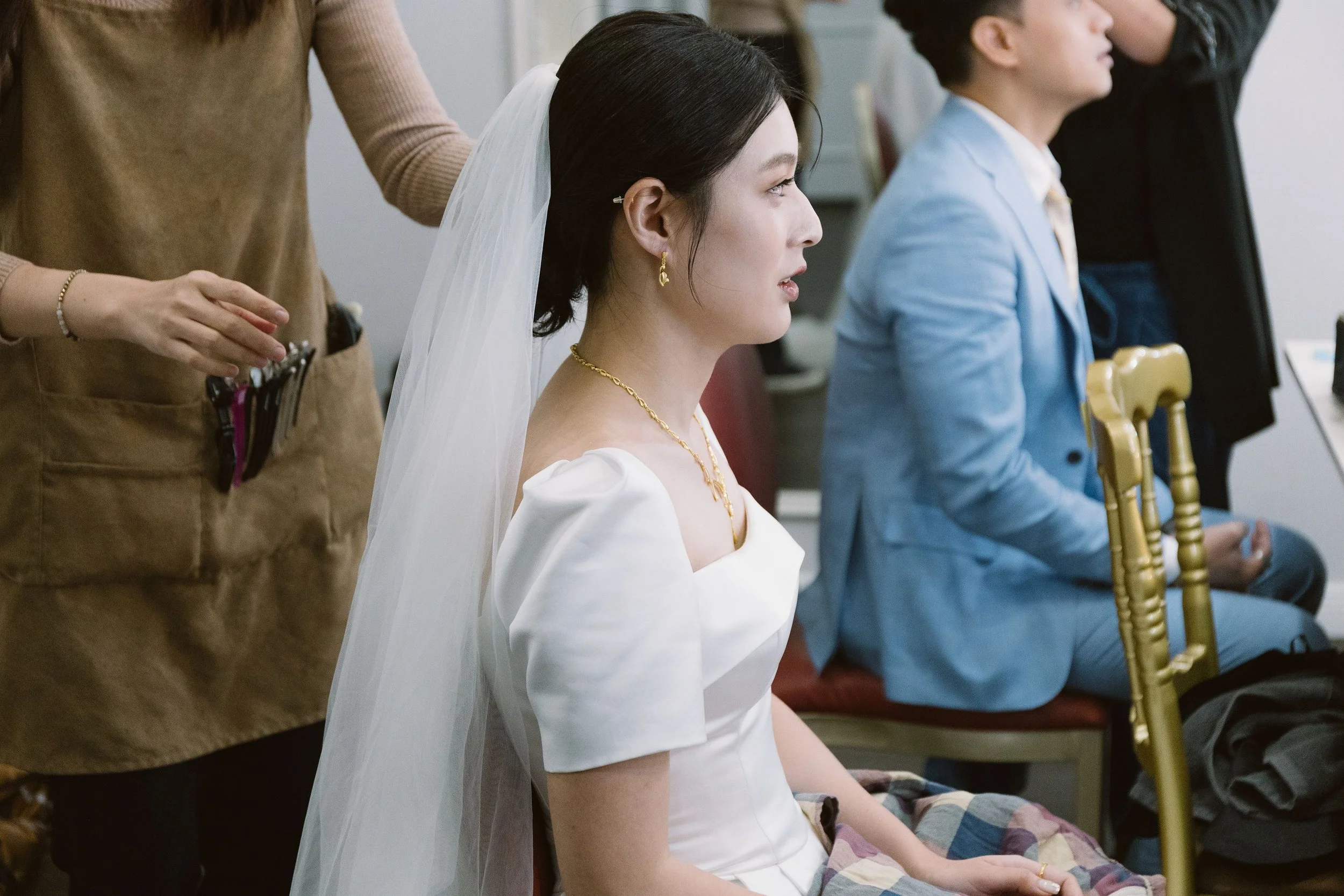 A woman in a wedding dress with a veil and gold jewelry is seated, with eyes closed, during a ceremony. A person standing behind her is adjusting her hair or veil. Other seated individuals are next to her, including a man in a light blue jacket, in a