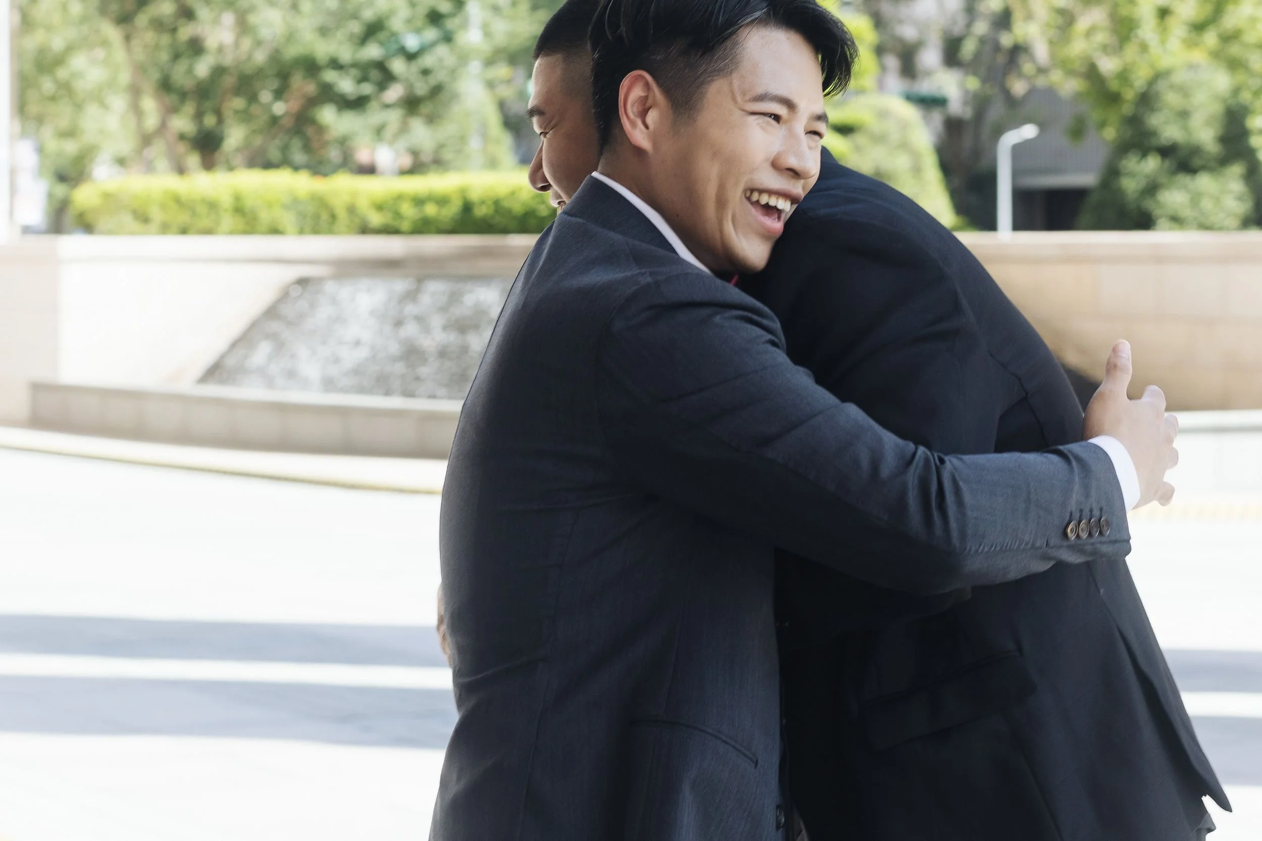 Two men in suits hugging and smiling outdoors with trees and a water feature in the background.