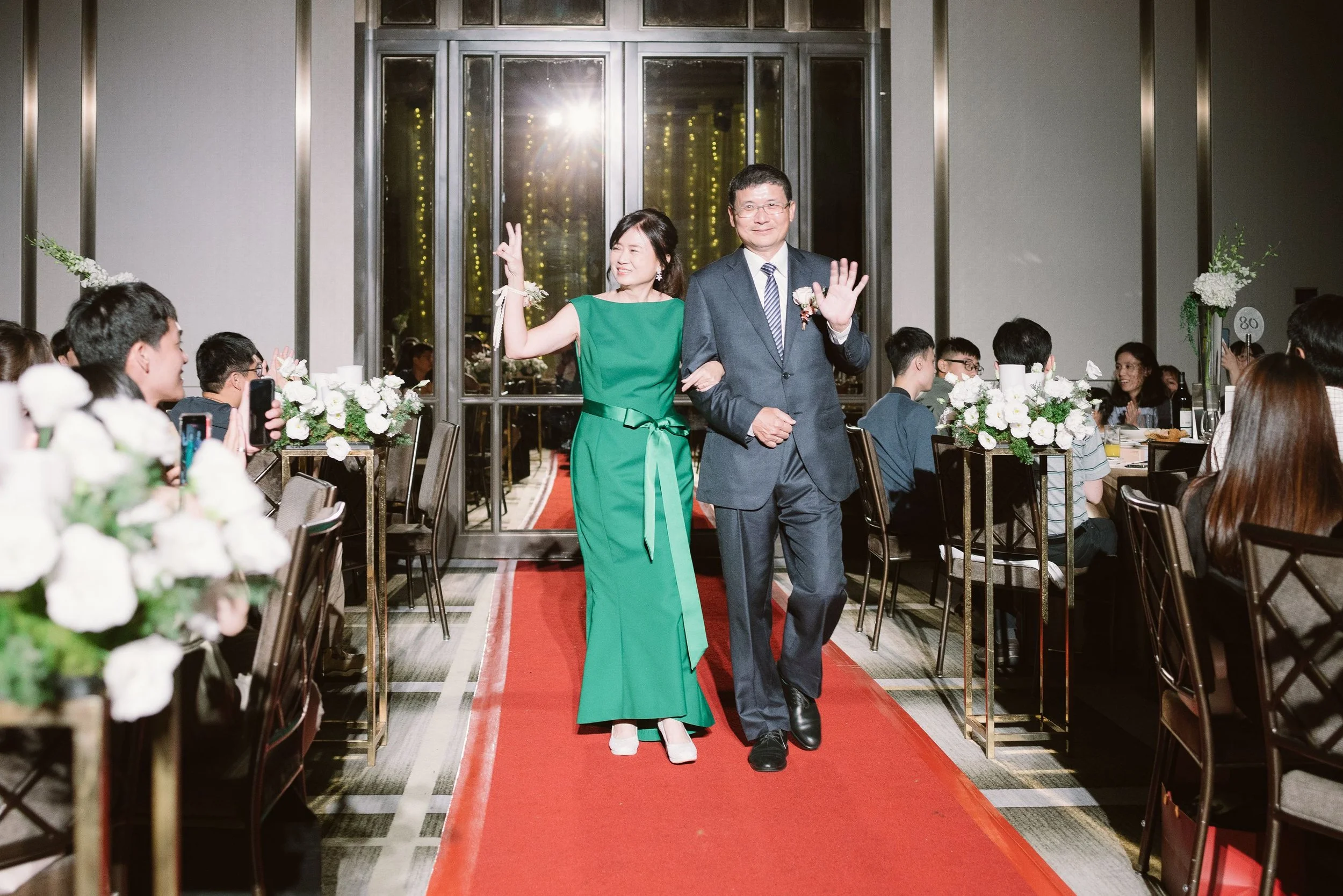 A woman in a green dress and a man in a gray suit walking down a red carpet at a formal event, surrounded by seated guests at decorated tables.