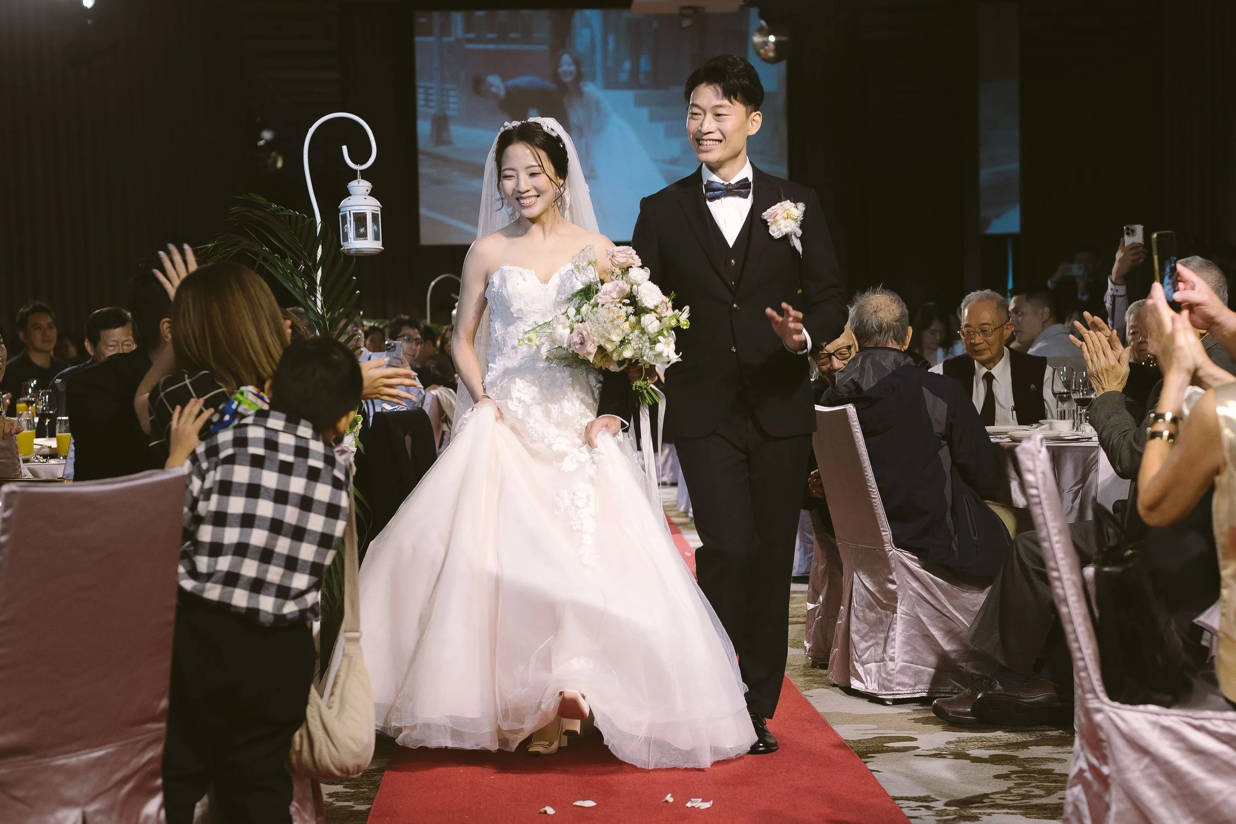 A bride and groom walking down the aisle at a wedding reception, smiling and holding flowers, surrounded by seated guests taking photos and clapping.