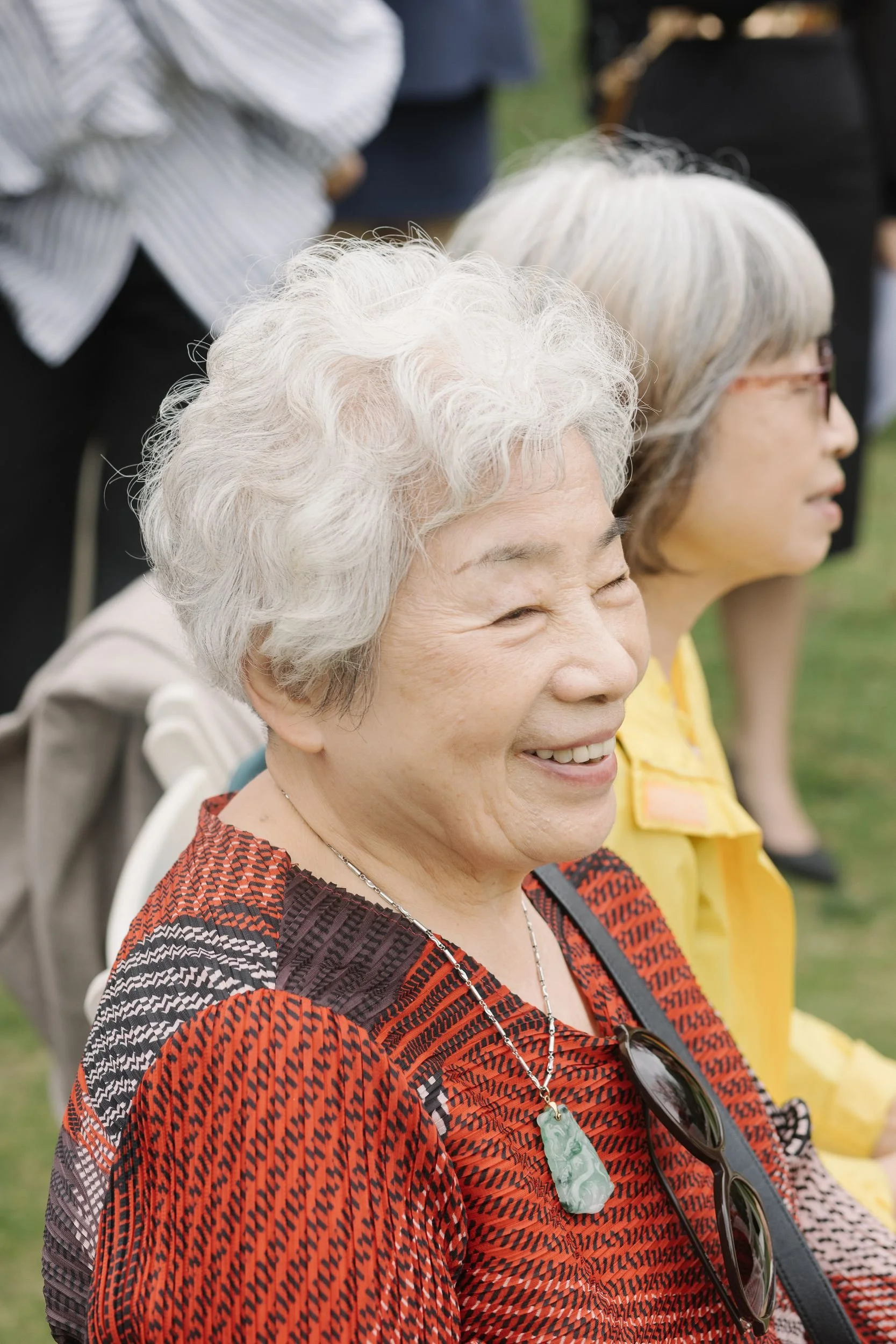 Smiling elderly woman with white curly hair, wearing a red and black patterned top, a green jade necklace, and sunglasses hanging from her shirt, sitting outdoors.