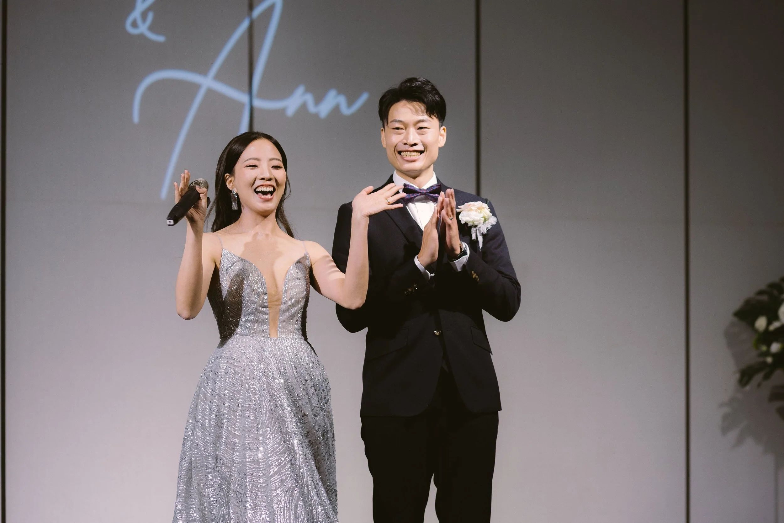 A smiling couple on stage at a wedding: the woman wears a silver, sparkly gown and holds a microphone, the man wears a black tuxedo with a bow tie and a white flower boutonniere. They are clapping and appear happy.