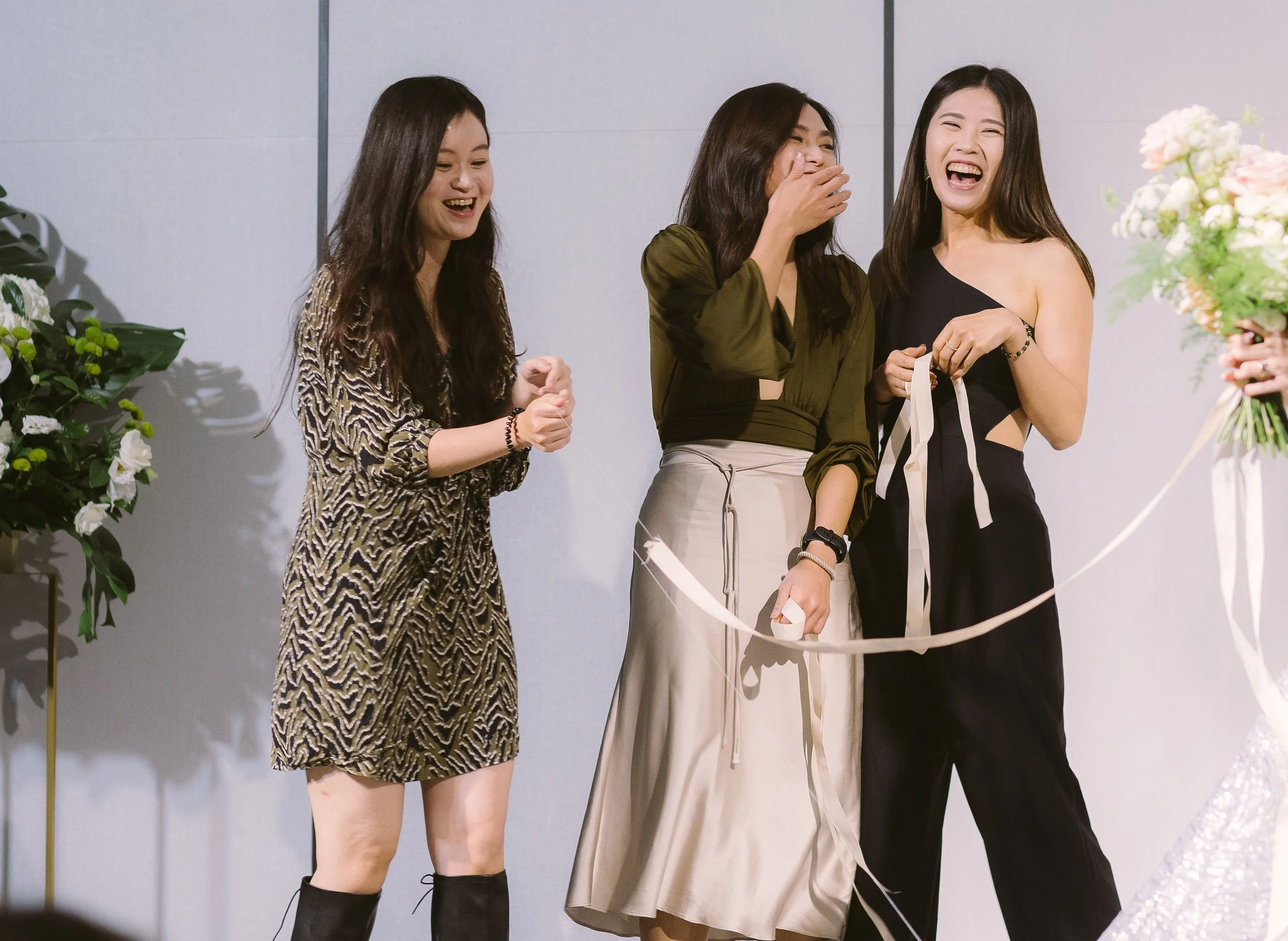 Three women standing together, laughing and smiling, during a social event. One woman is holding a ribbon, with flowers nearby.