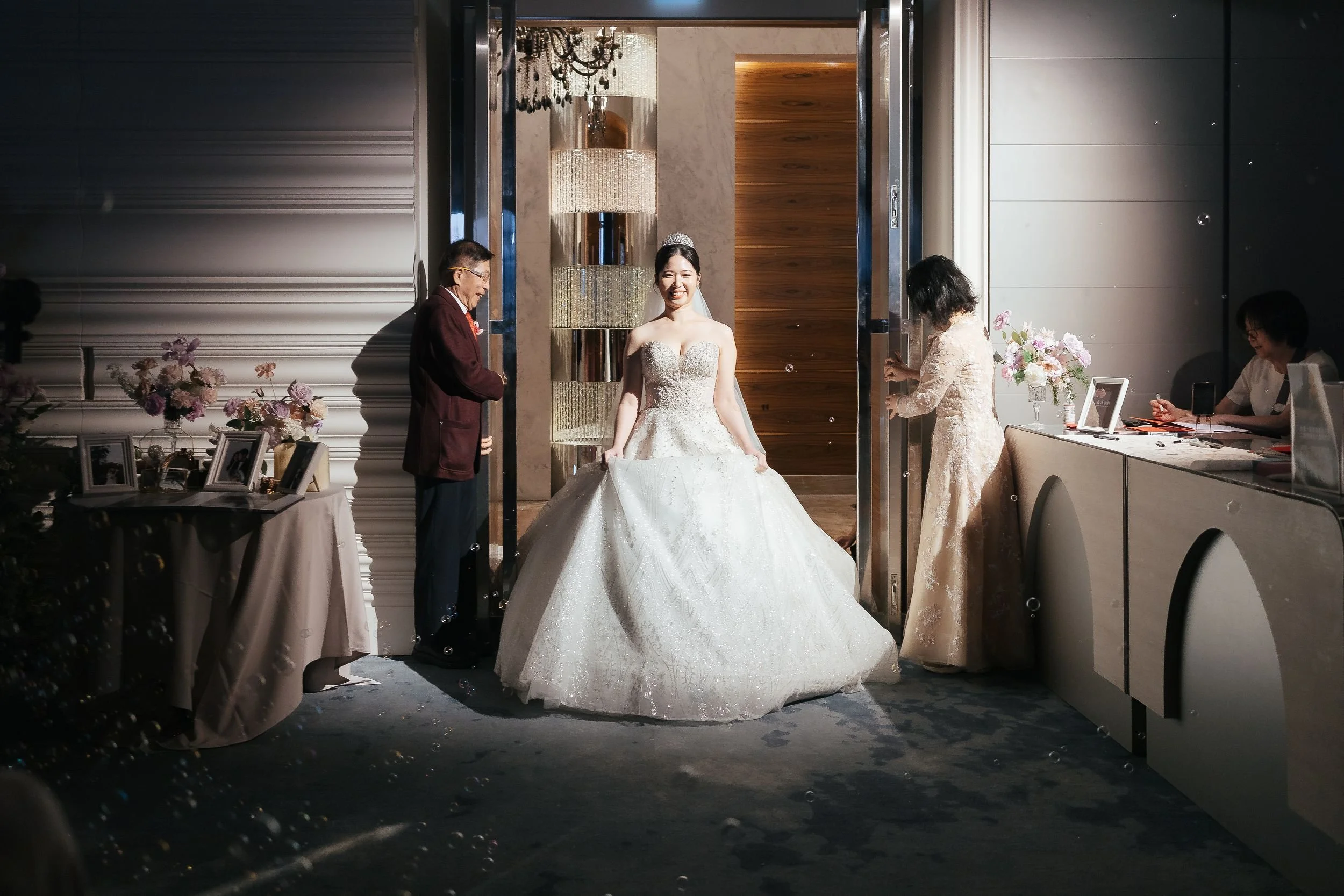 A bride in a strapless wedding gown and tiara is smiling as she stands in an elevator, surrounded by wedding guests at a celebration with decorated tables, flowers, and framed photos.