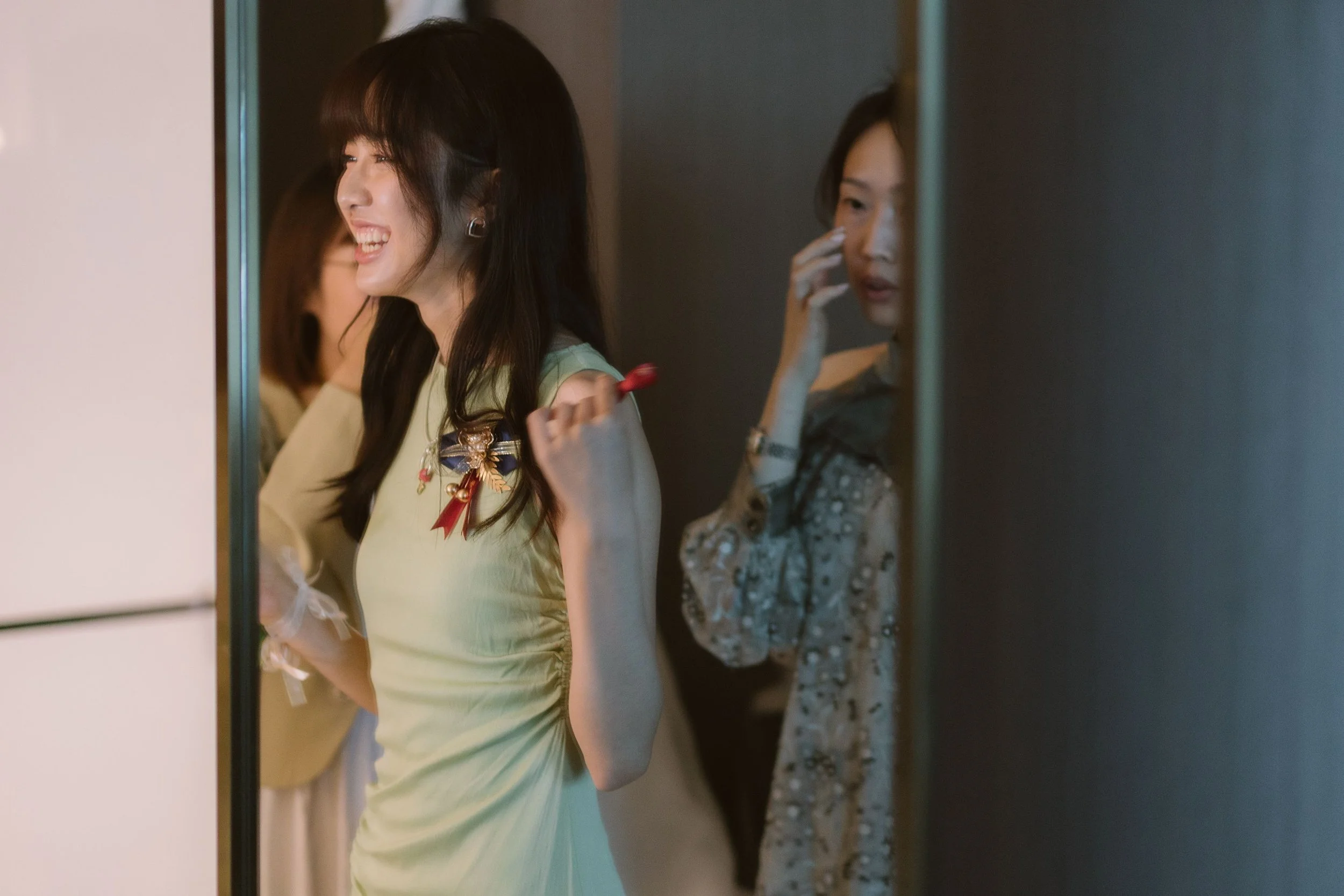 Two women smiling, one in a cream dress with a decorative bow and brooch, and the other in a patterned dress, standing behind a mirror.