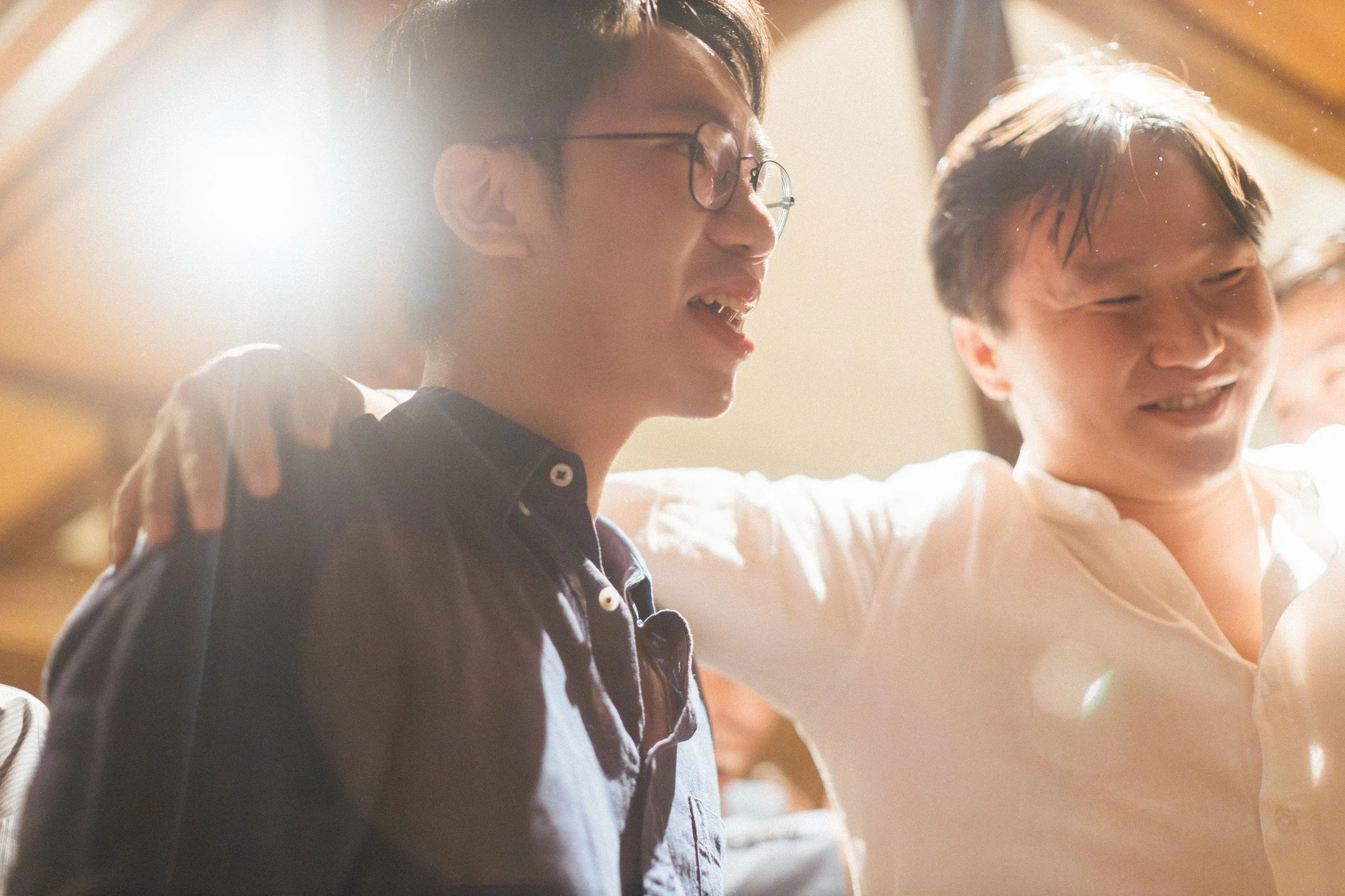 Two young men are smiling and embracing each other, with the person on the left wearing glasses and a dark shirt. The sunlight is shining warmly, creating a soft, joyful atmosphere.