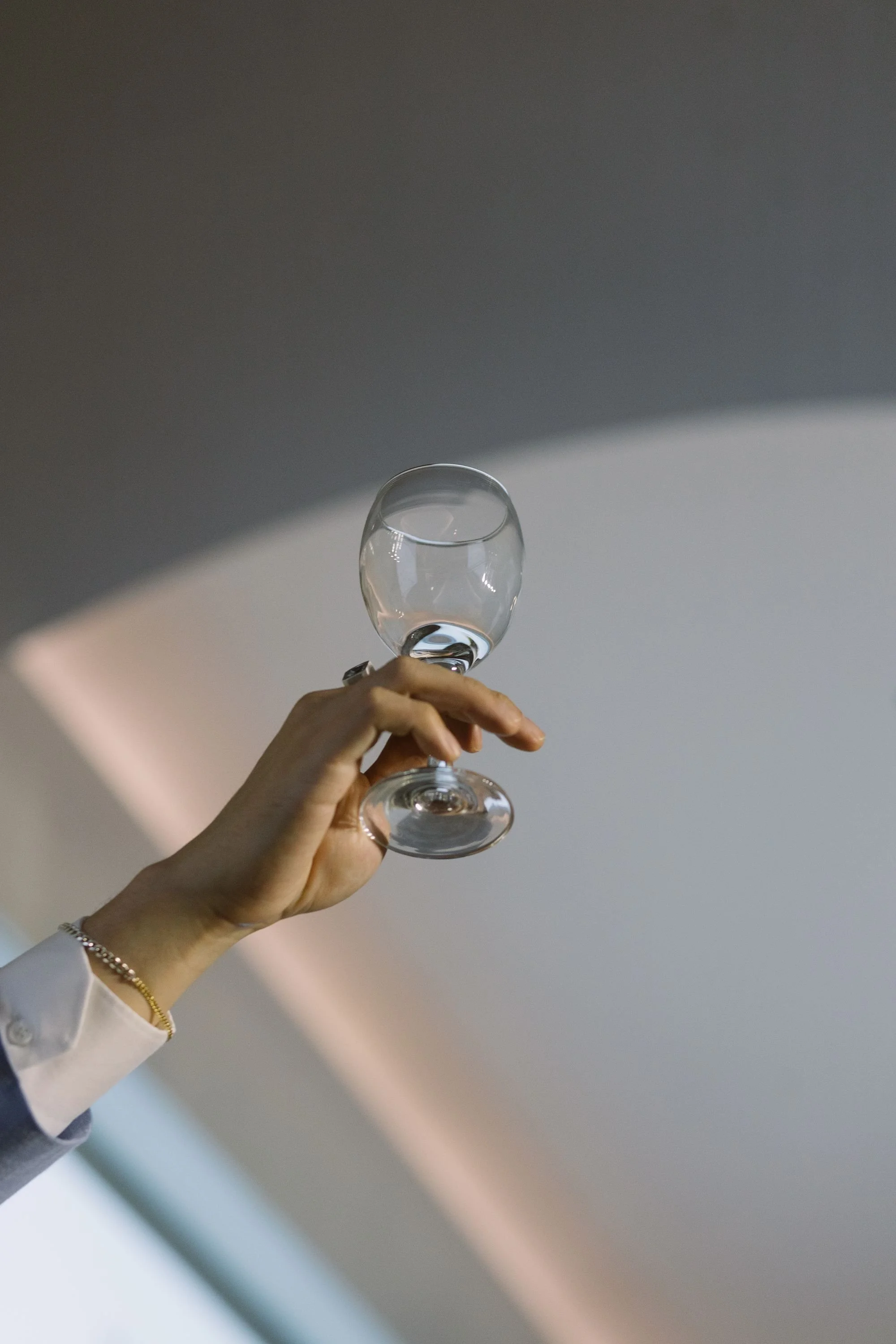 A person holding an empty wine glass against a curved wall with soft lighting. The person's hand is visible, wearing a gold bracelet and white or light-colored sleeve.