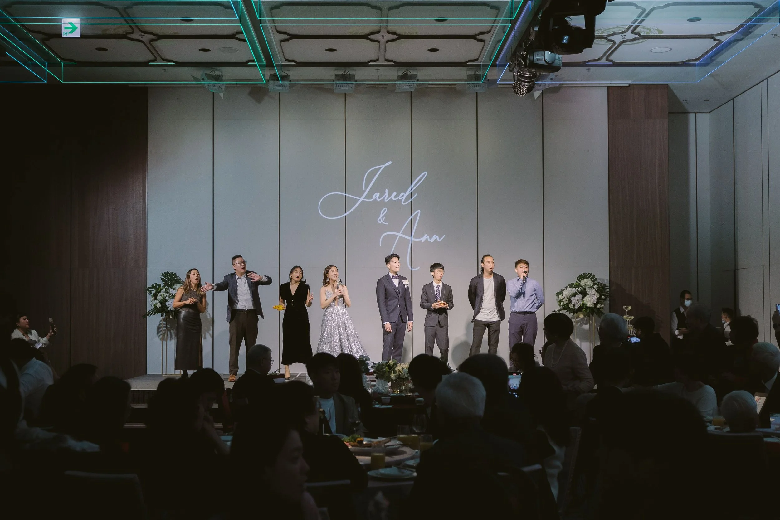 A group of people dressed in formal attire standing on a stage at a wedding reception. The background has a large sign with the names Jarad and Ann. There are floral arrangements on either side of the stage and an audience seated in front.