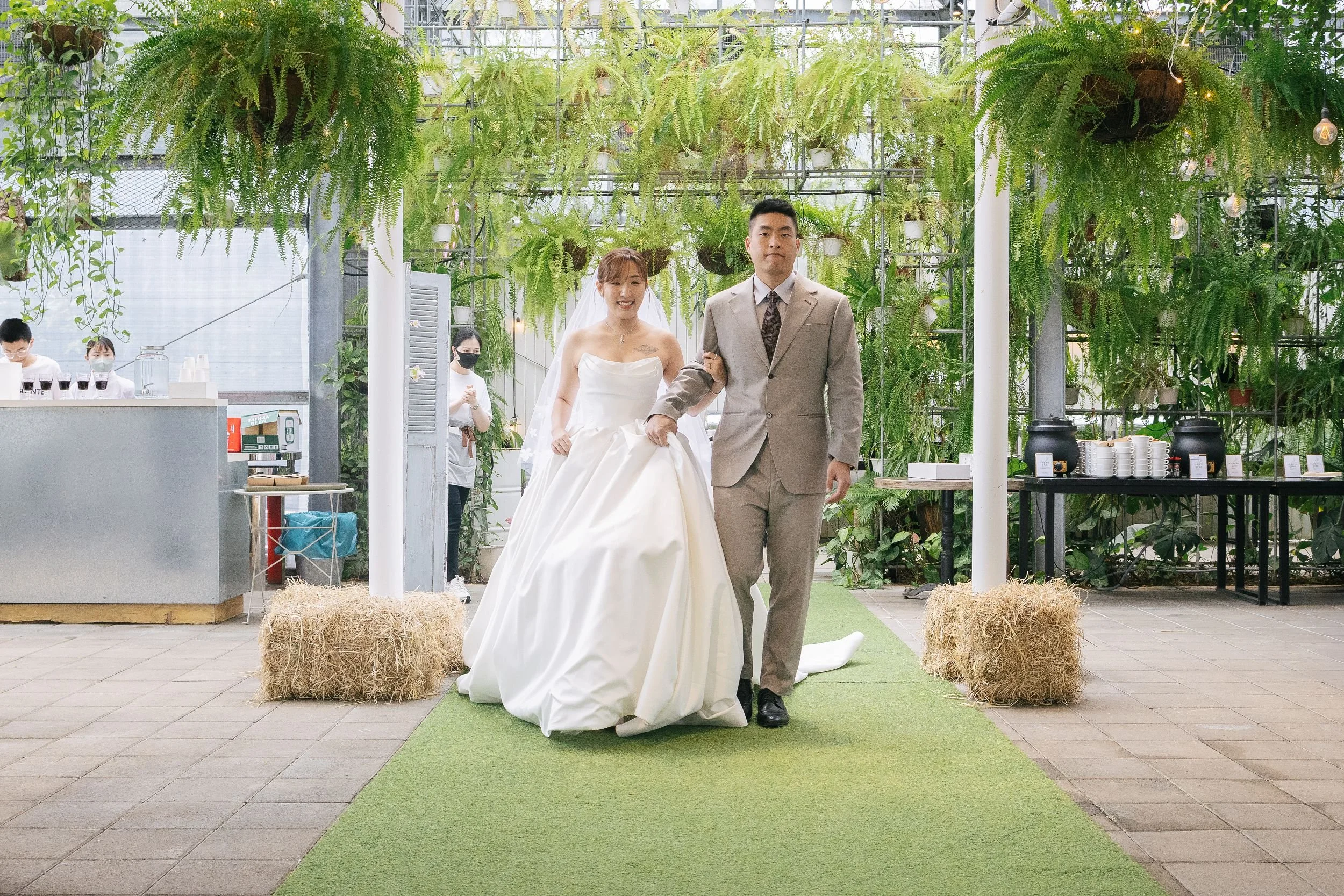 A bride and groom walking arm-in-arm on a green aisle in a greenhouse. The bride is wearing a strapless white wedding gown and veil, with a necklace, smiling. The groom is wearing a beige suit with a patterned tie, smiling slightly. Behind them are g