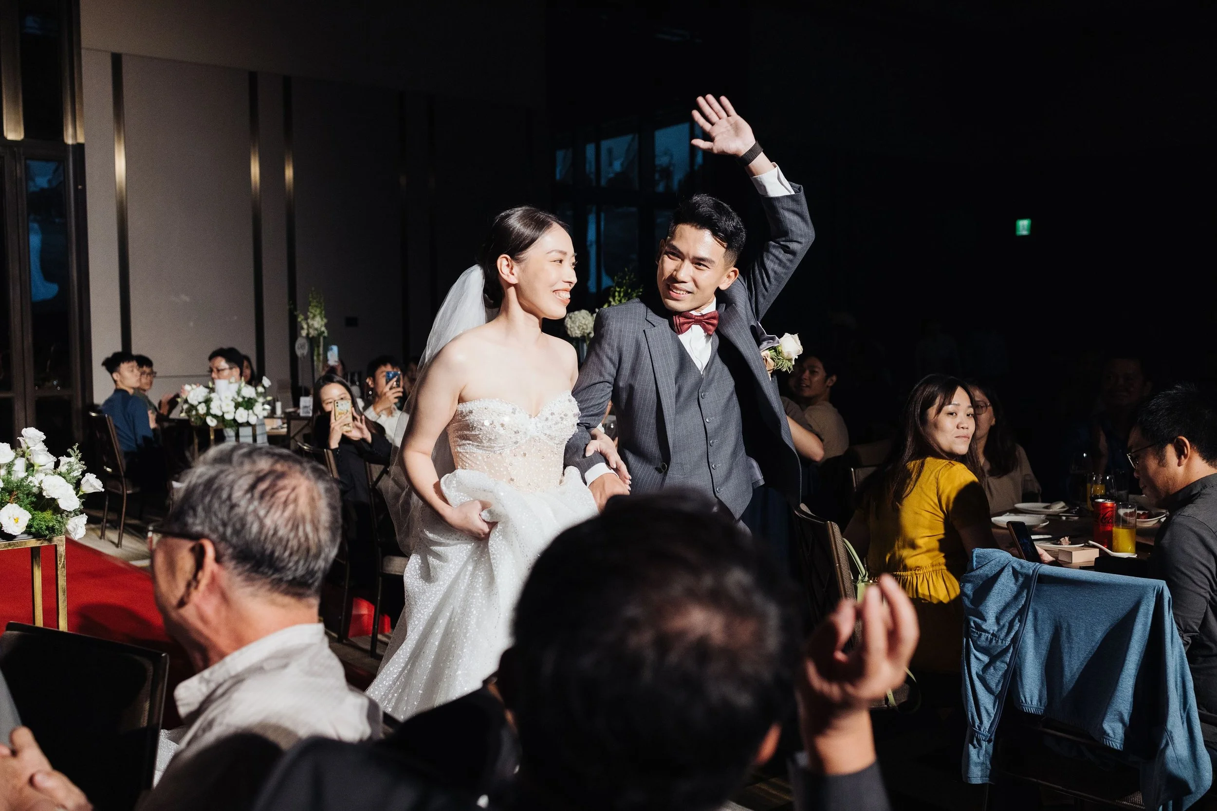 A bride and groom at their wedding reception, surrounded by seated guests, with the groom raising his arm and the bride smiling.