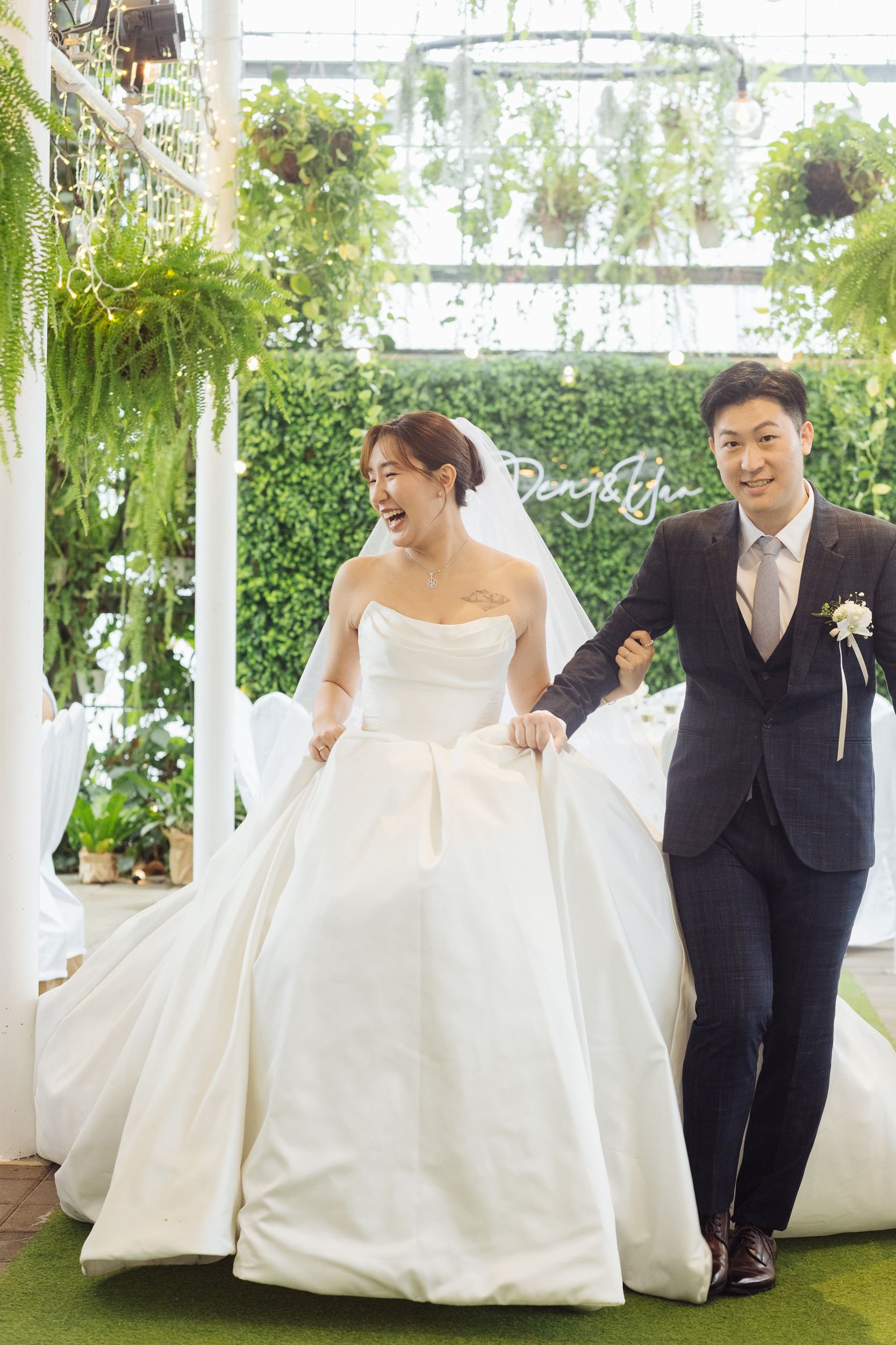 A happy bride in a white wedding dress and a groom in a dark suit holding hands indoors with green plants and hanging lights in the background.
