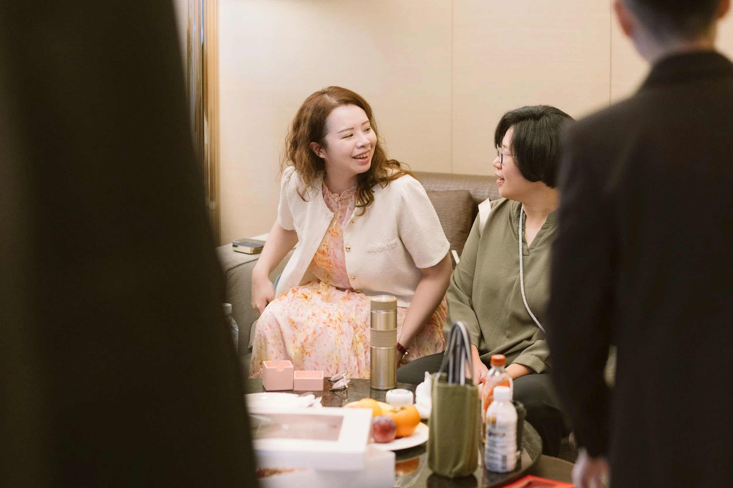 Two women are sitting at a table, engaged in conversation during a social gathering or event in an indoor setting. The table has various objects on it, including bottles and personal items.