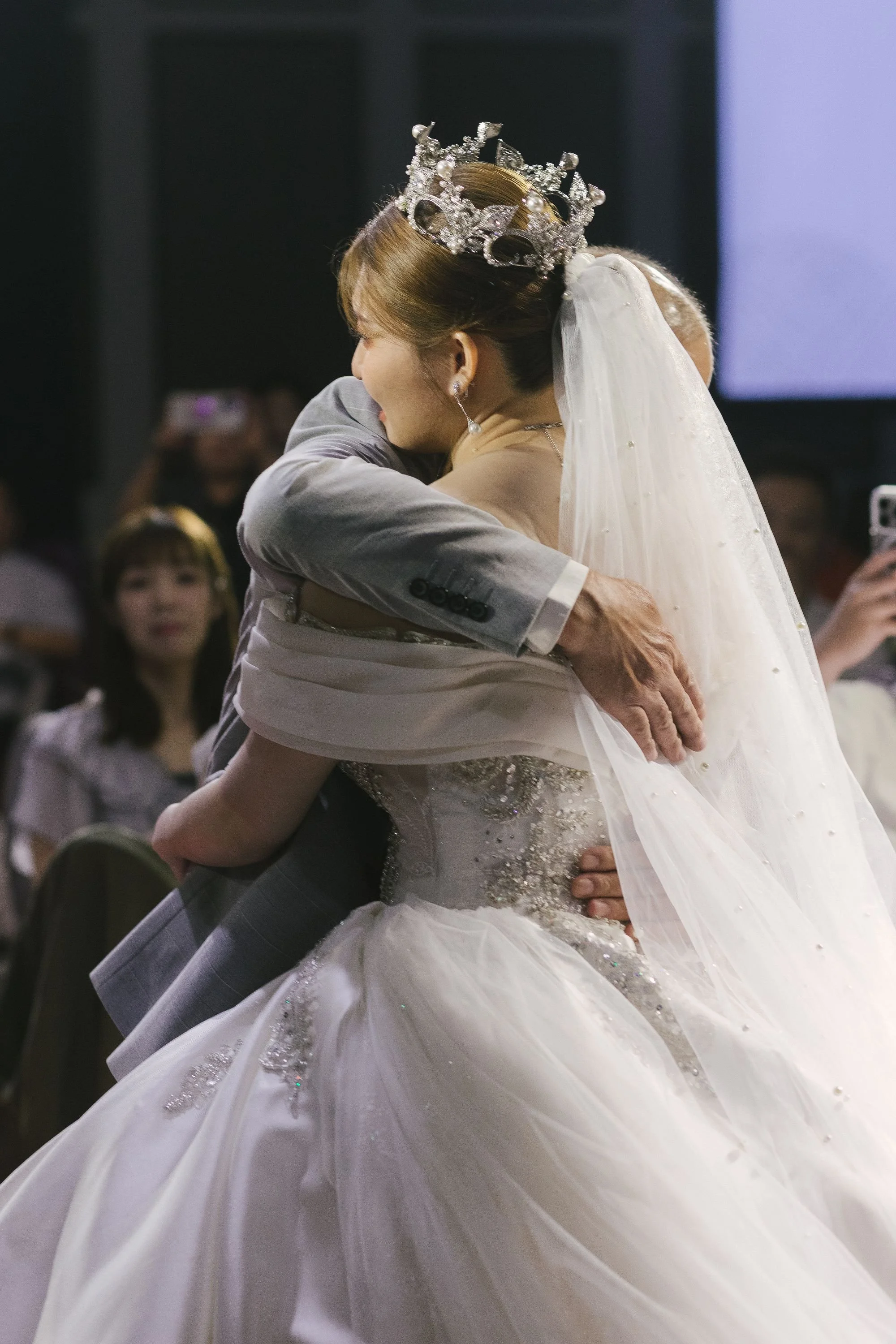 A bride and groom hugging at a wedding reception, with the bride wearing a tiara and veil, and the groom dressed in a suit.