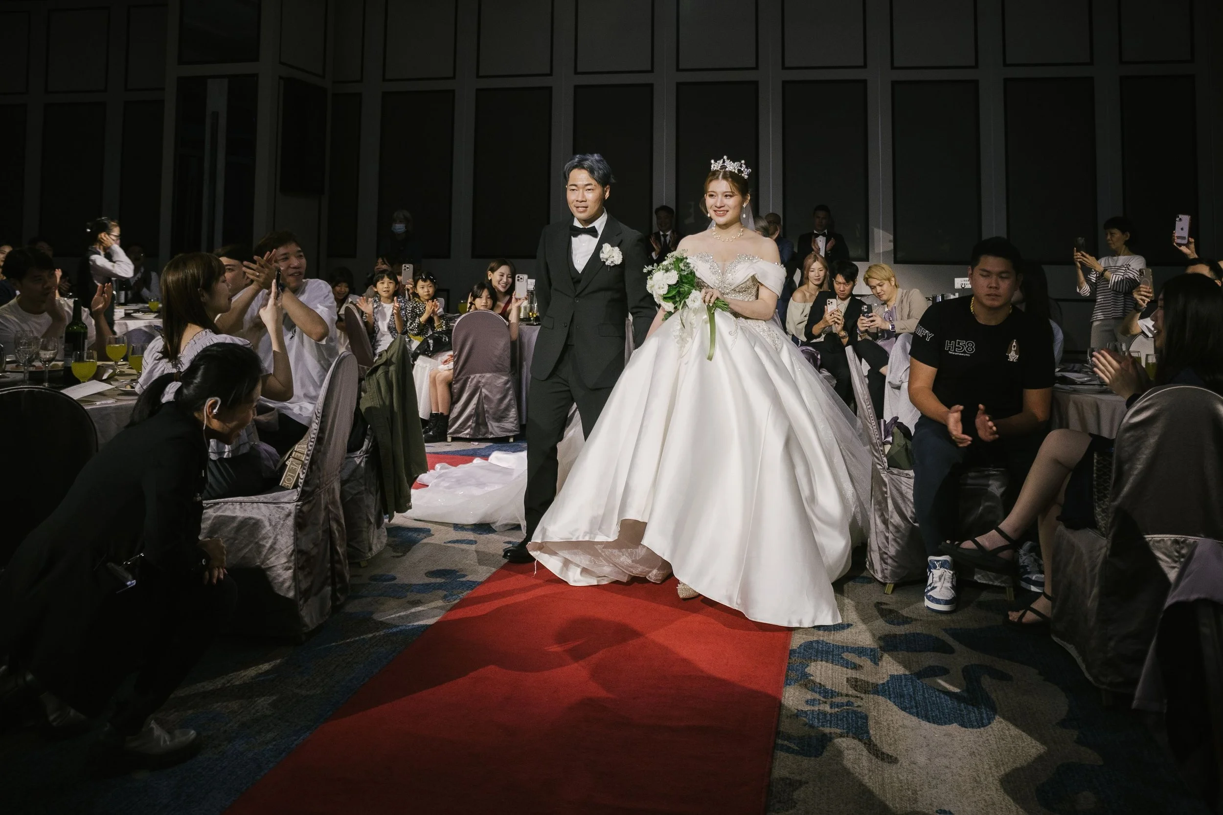 A bride and groom walking down a red carpet at their wedding reception, surrounded by guests taking photos and cheering.