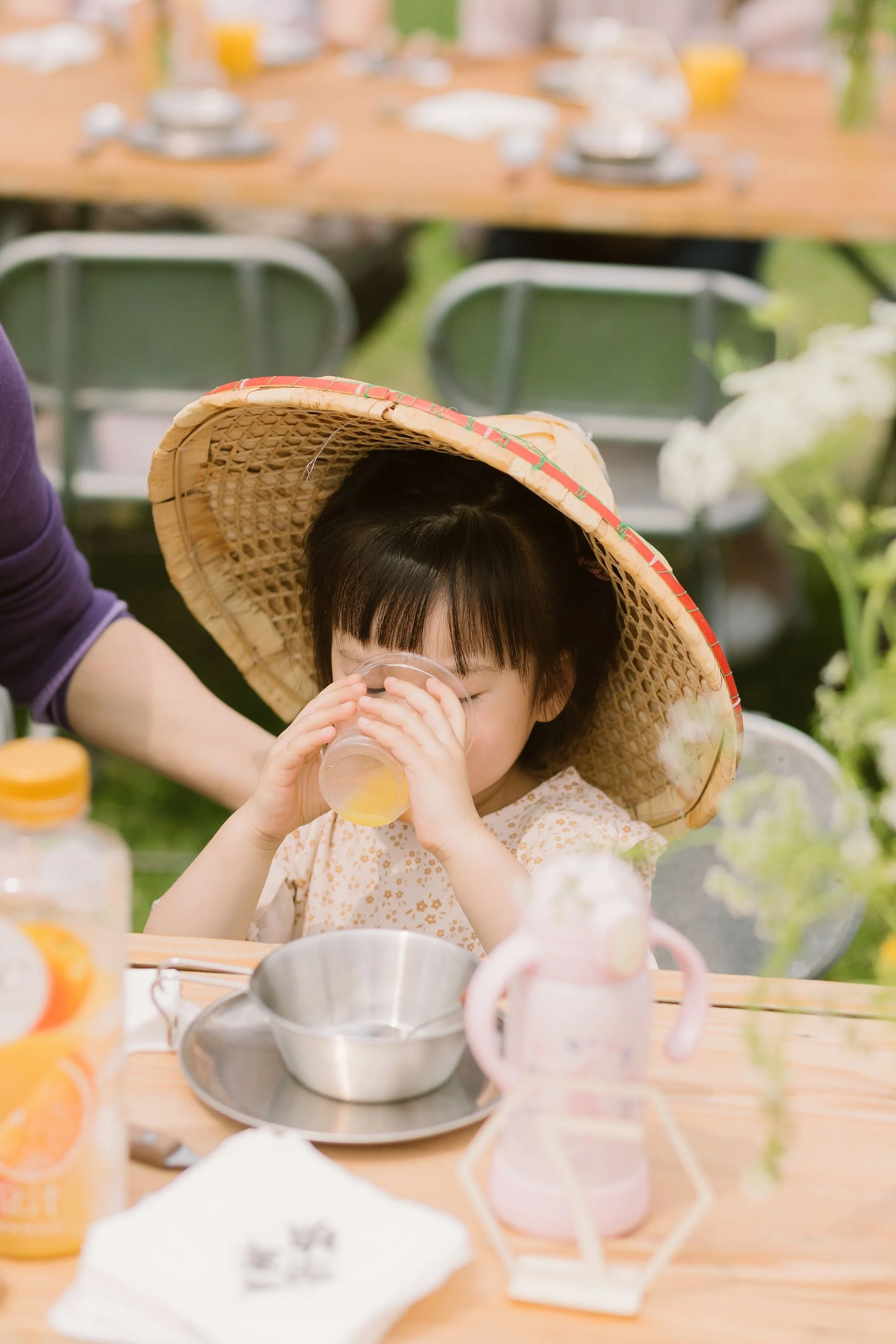 A young girl wearing a wide-brimmed straw hat and a light-colored dress is drinking from a clear glass at an outdoor gathering. The background shows a blurred table set with plates, glasses, and cups, indicating a festive occasion.