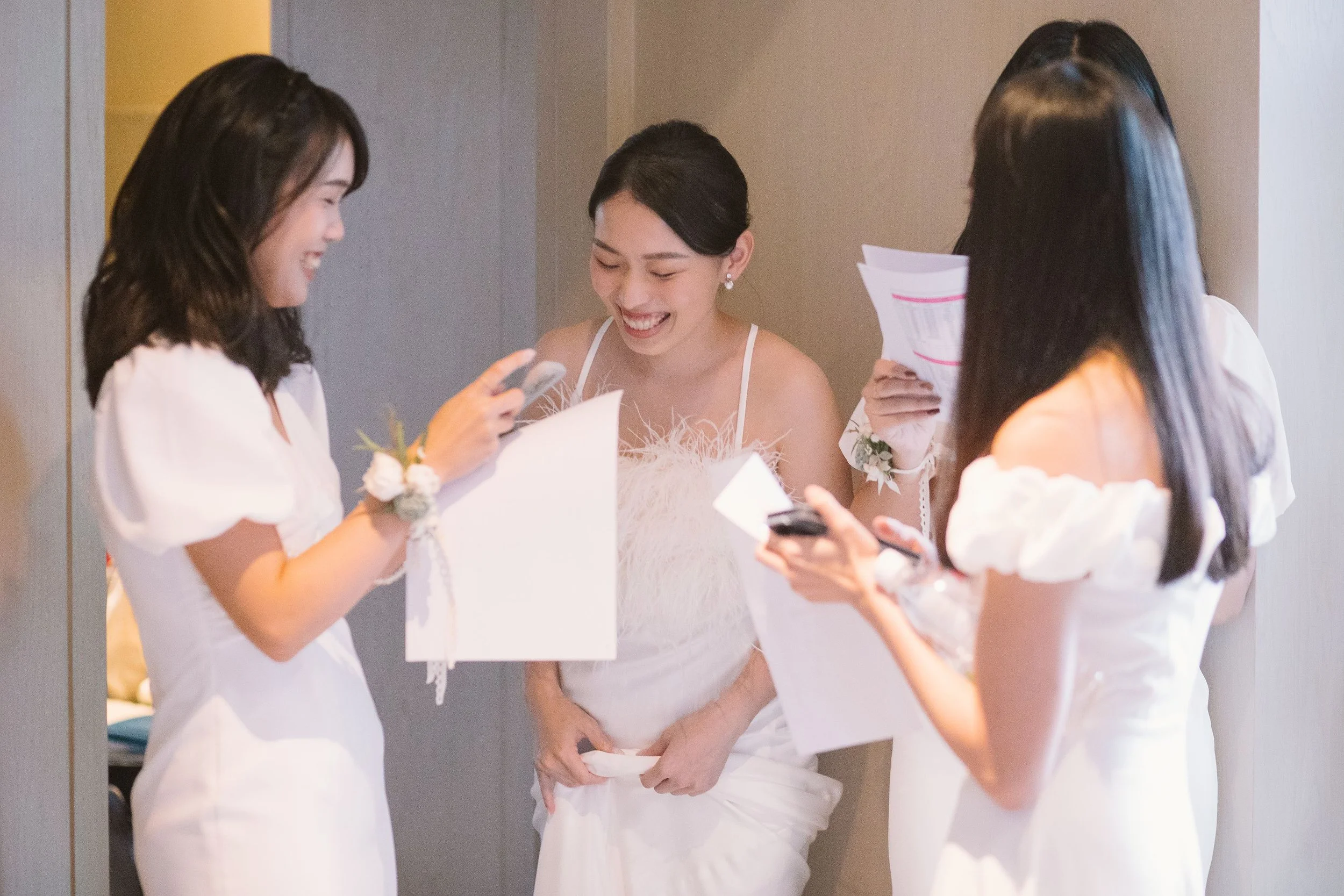 Four women dressed in white are standing together, smiling, and looking at their phones and papers in a brightly lit room.