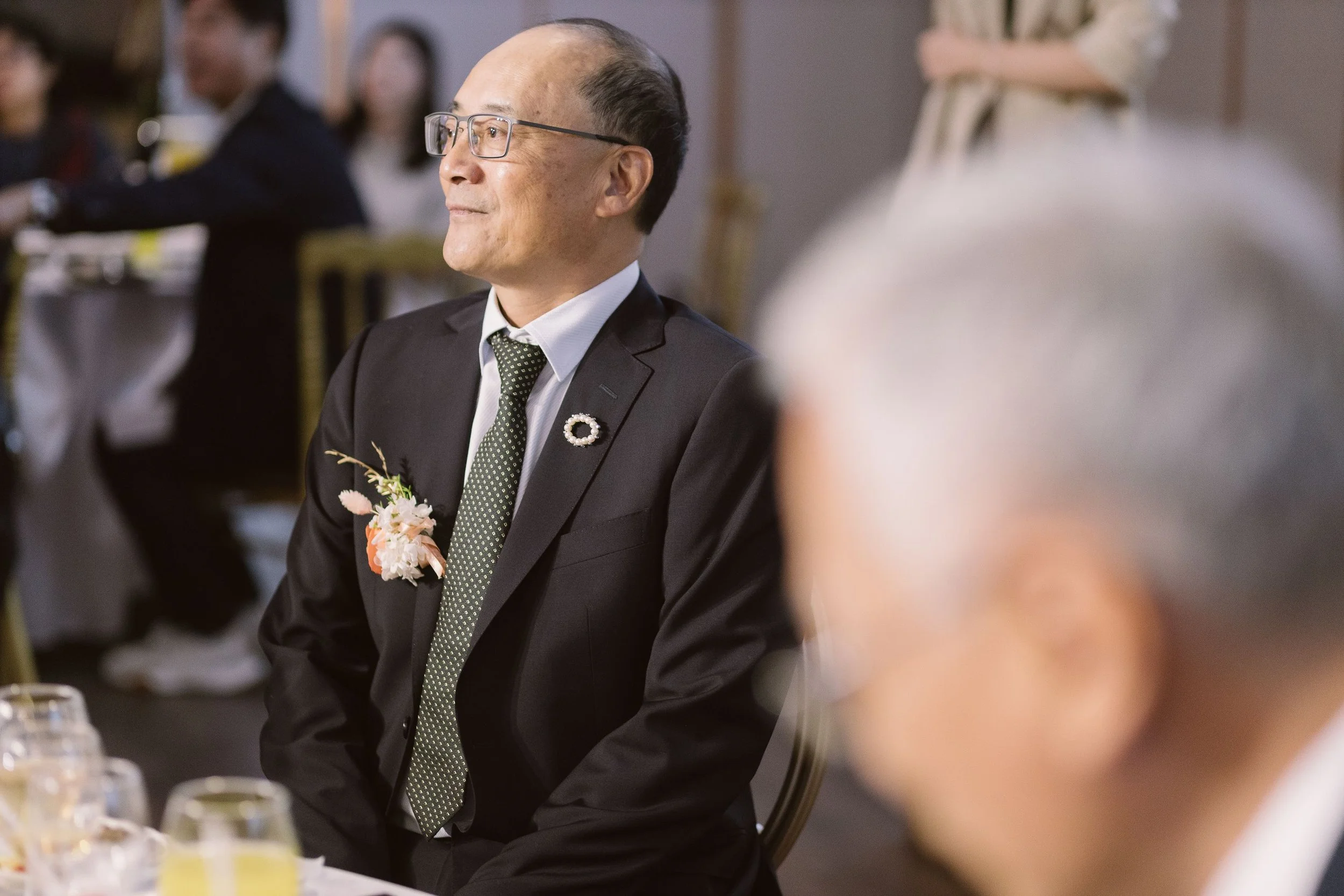 An elderly man wearing glasses, a dark suit, white shirt, and a green patterned tie, sits at a table during a social gathering. He has a floral boutonniere pinned to his suit lapel.