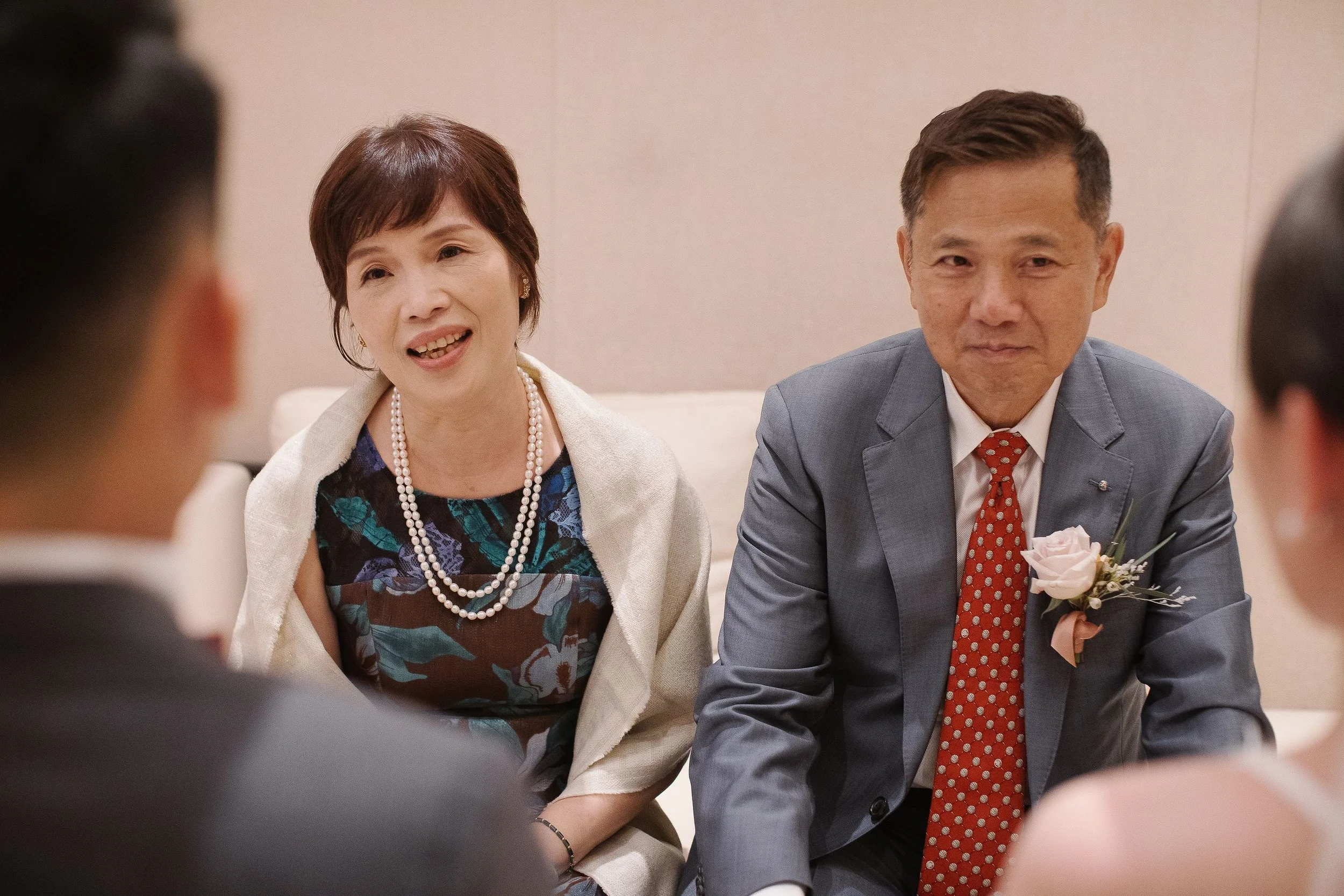 A woman with short brown hair wearing a pearl necklace and patterned dress, and a man with short hair wearing a suit and red polka dot tie, sitting at a table.