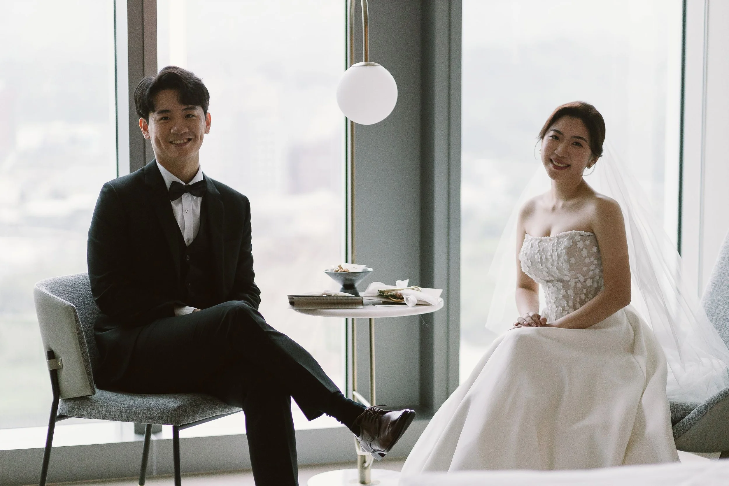 A bride and groom sitting indoors near large windows, smiling at the camera. The groom is wearing a black tuxedo with a bowtie, and the bride is in a strapless wedding gown with a veil. They are seated at a small table with some items on it, includin