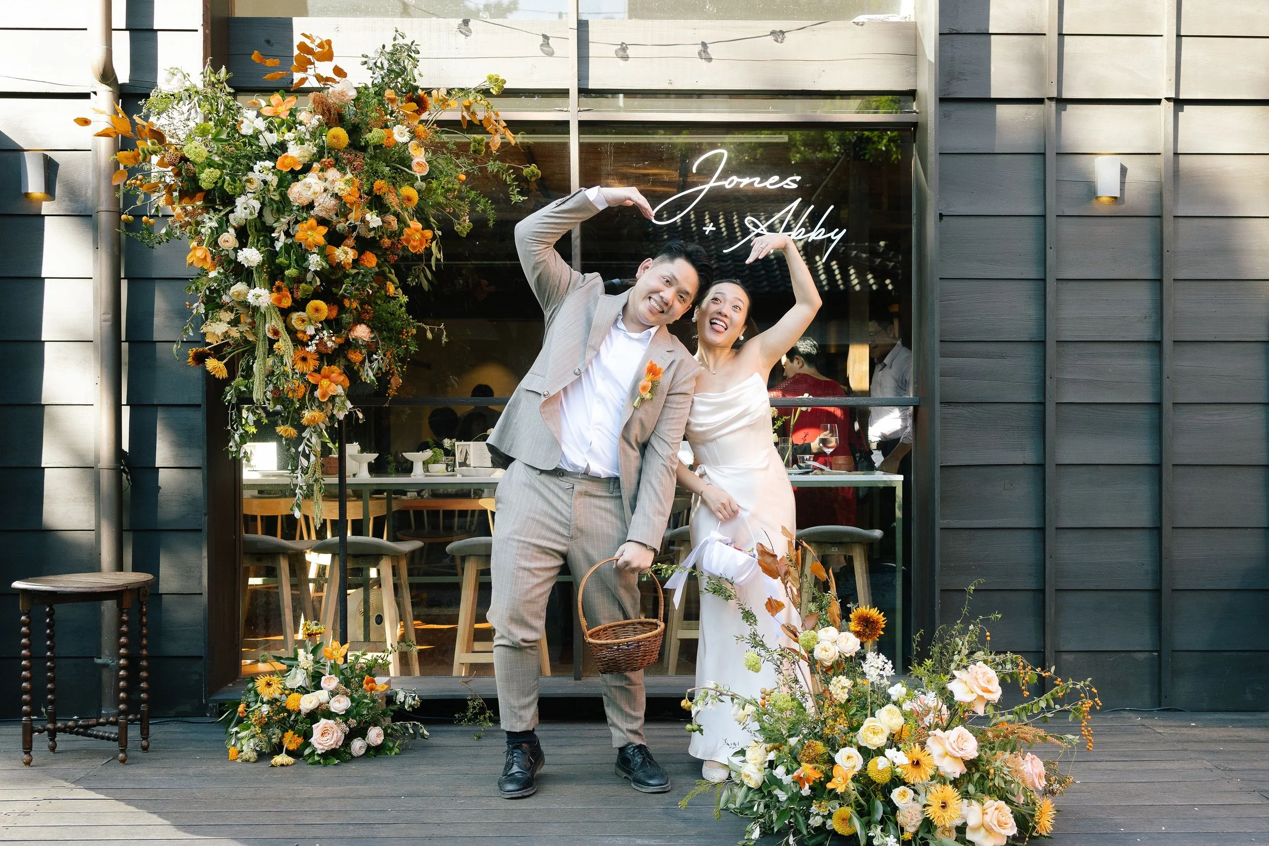 A couple at a wedding or celebration standing in front of a decorated outdoor space with flowers. The man is in a light gray suit and the woman in a white dress, both smiling and making heart shapes with their arms over their heads. The background fe