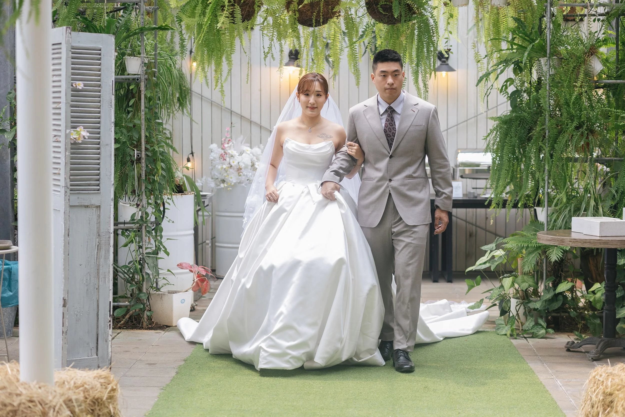 A bride in a white wedding gown and a groom in a gray suit walk together down an aisle decorated with hanging plants and greenery in an indoor garden or greenhouse setting.