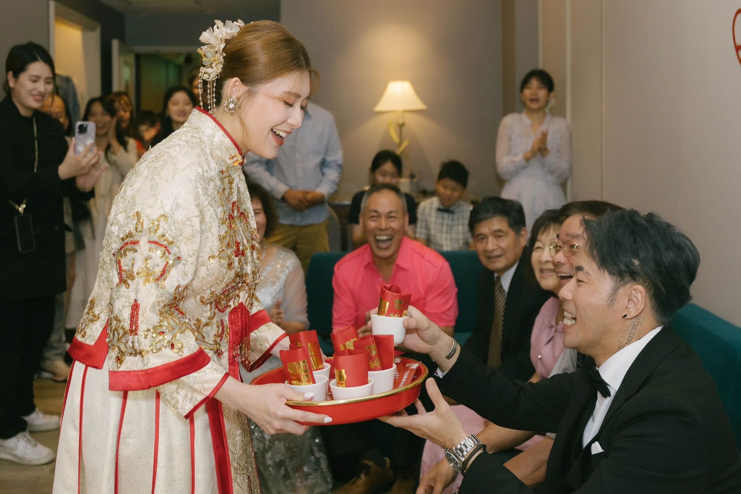 A woman dressed in traditional Chinese wedding attire presents red envelopes to a man at a celebratory gathering. The group of people around her are smiling and engaging in the exchange, with some holding red packets and others capturing the moment o