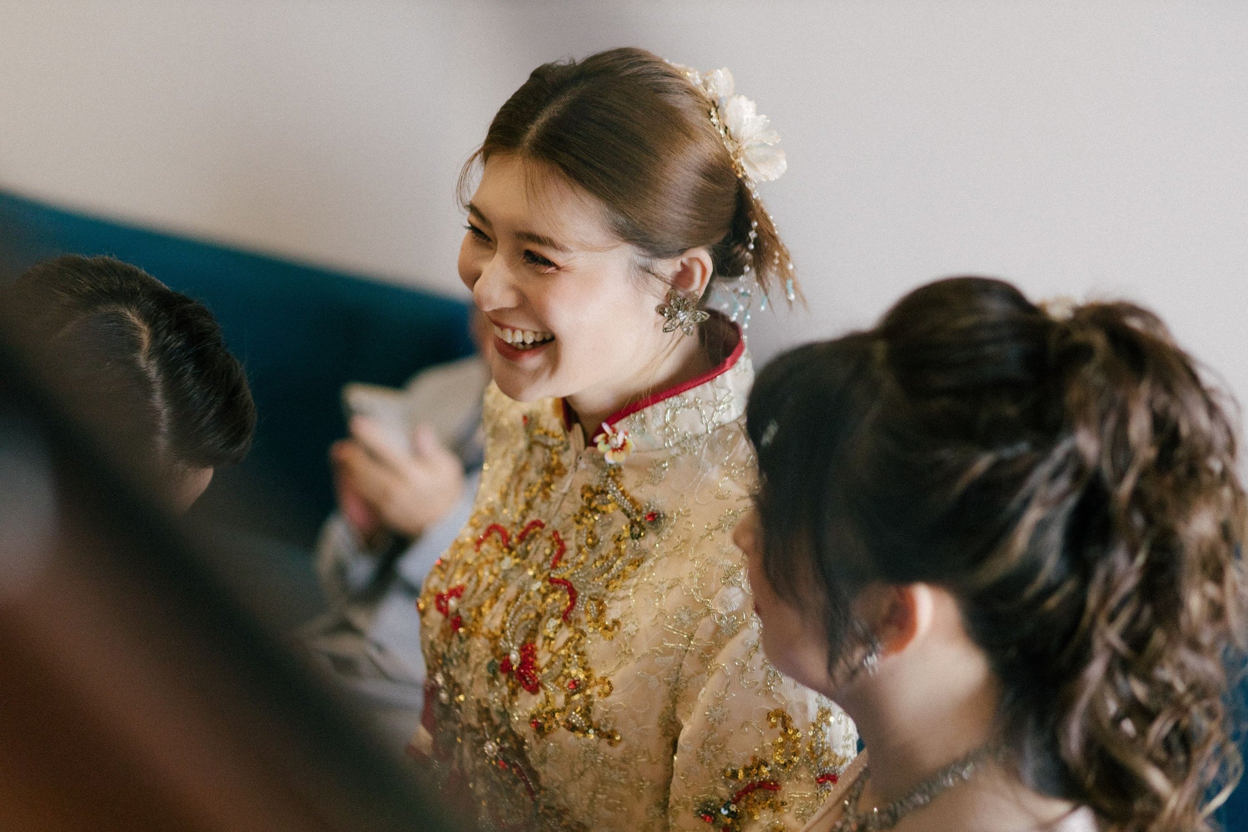 A woman dressed in an ornate, embroidered dress with gold and red details, smiling and looking to her right, surrounded by other women with dark hair, in an indoor setting.