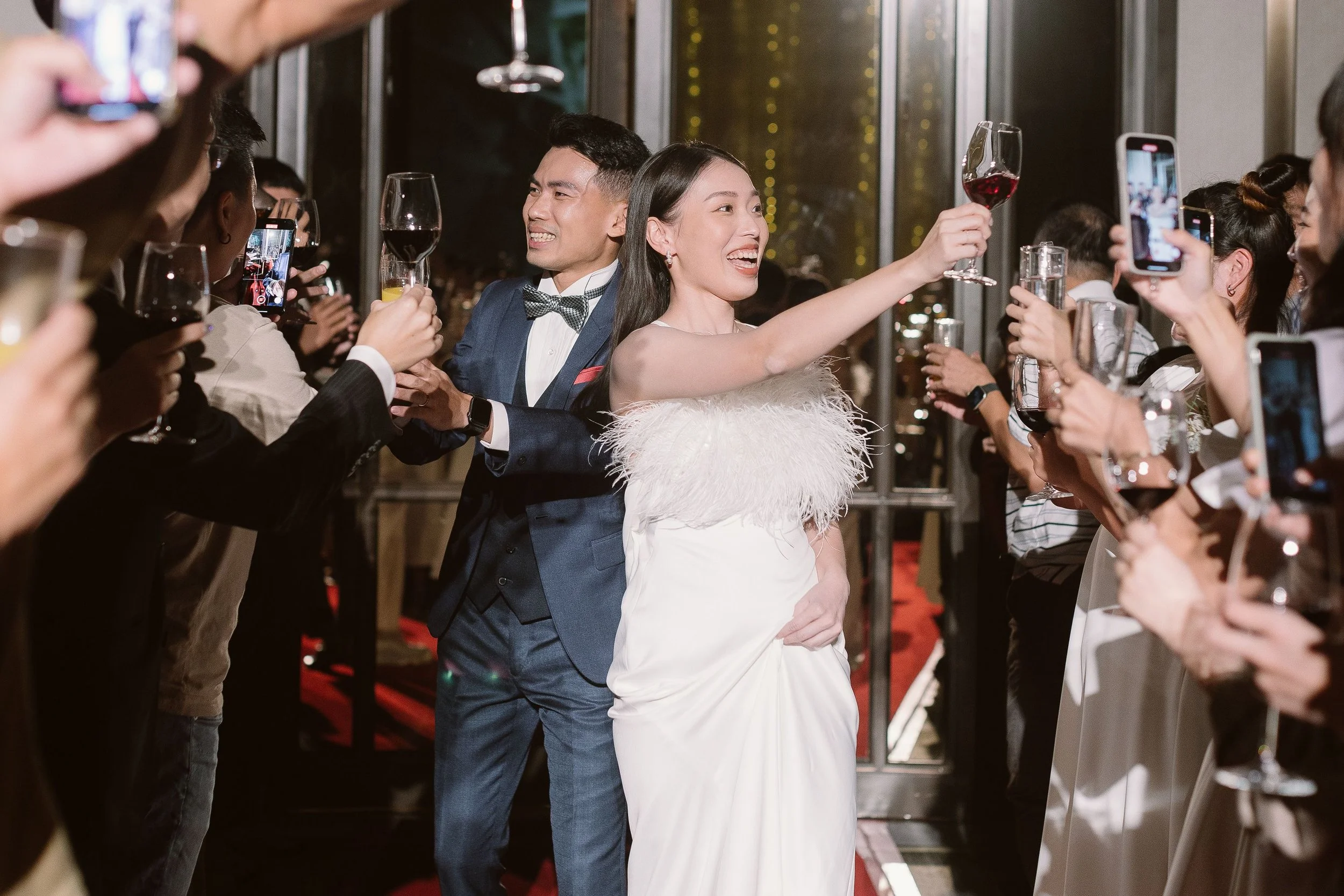 A bride and groom smile and toast with glasses of wine at their wedding reception. Guests around them hold cameras and phones to capture the moment.