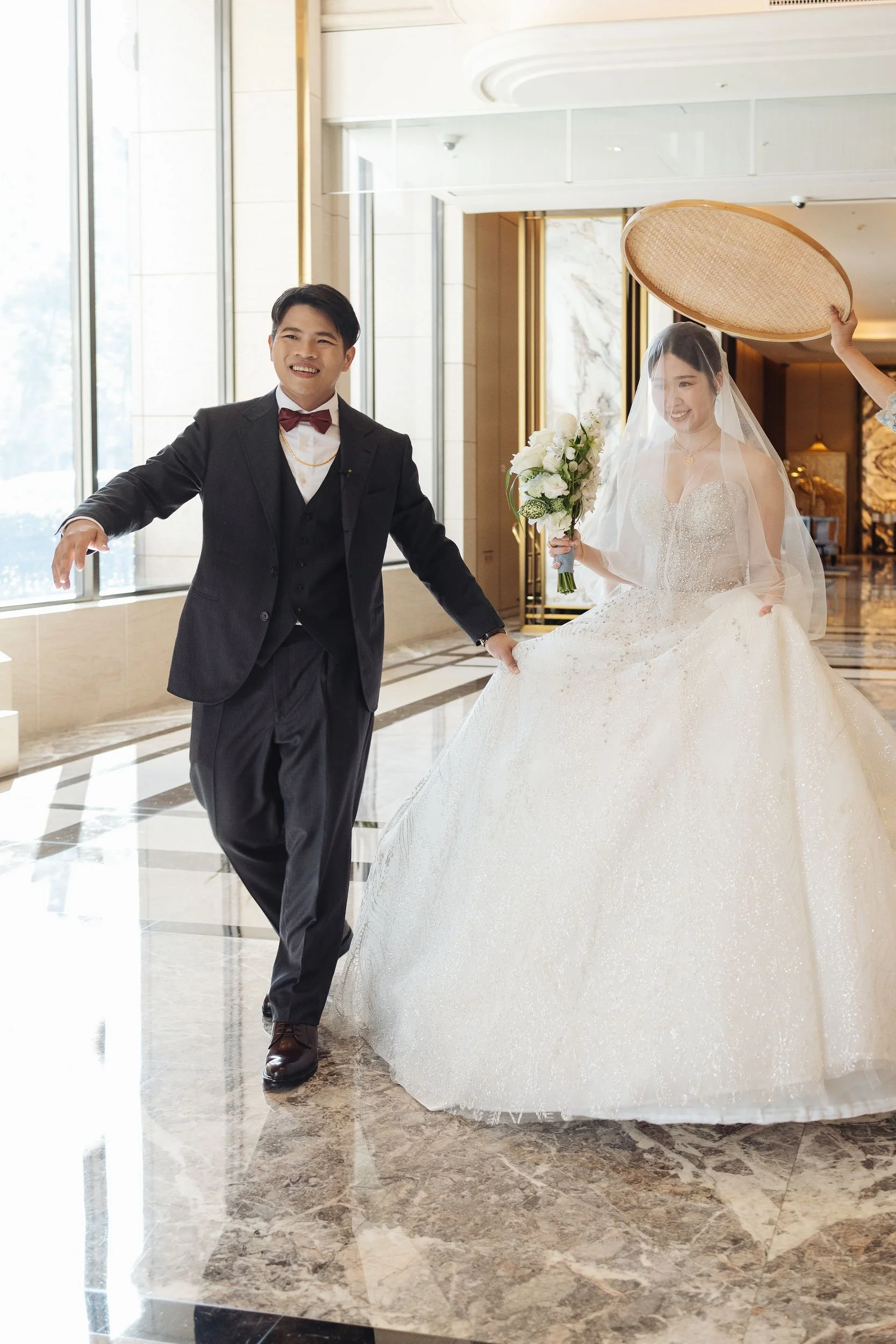 A bride in a wedding dress holding a bouquet and a groom in a black suit with a red bow tie standing inside a luxurious building, with an attendant holding a round tray above the bride's head.
