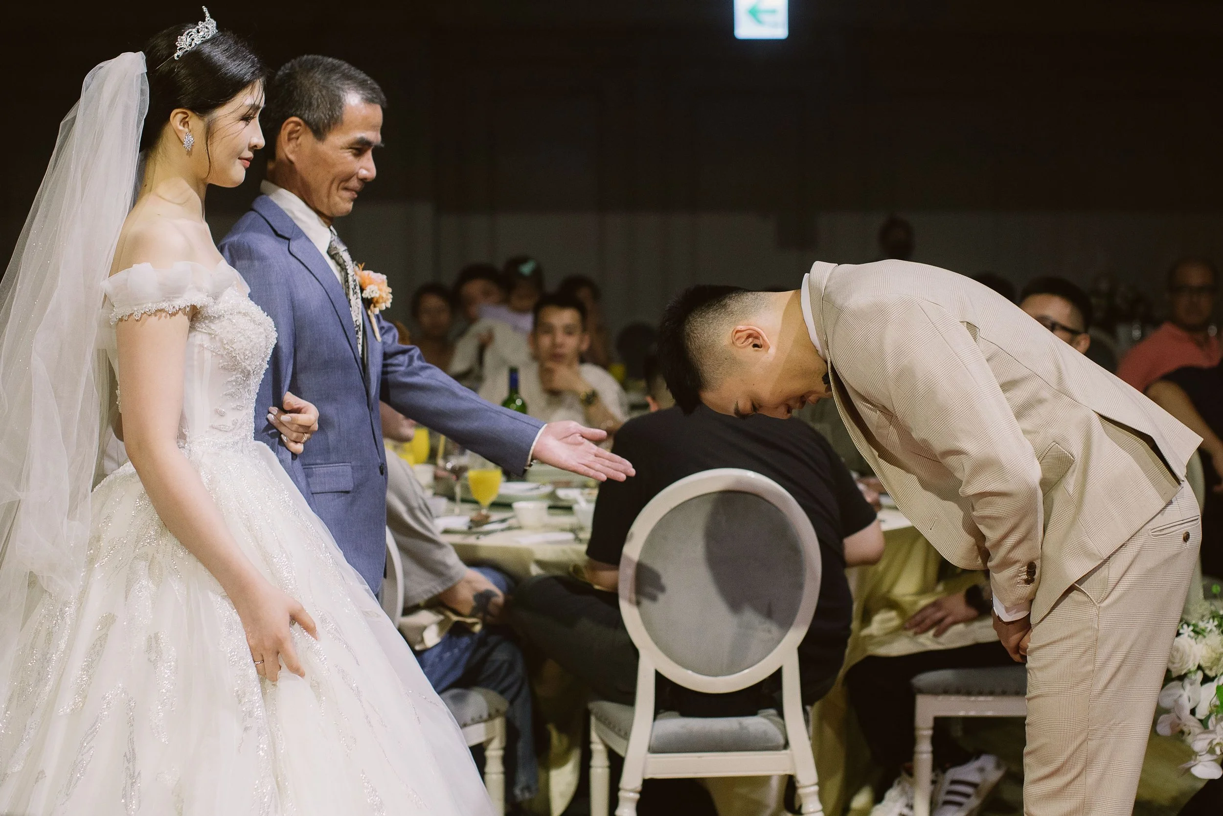 A bride in a wedding dress and tiara, standing next to a man in a suit, with a woman bowing in front of them during a wedding ceremony surrounded by guests.