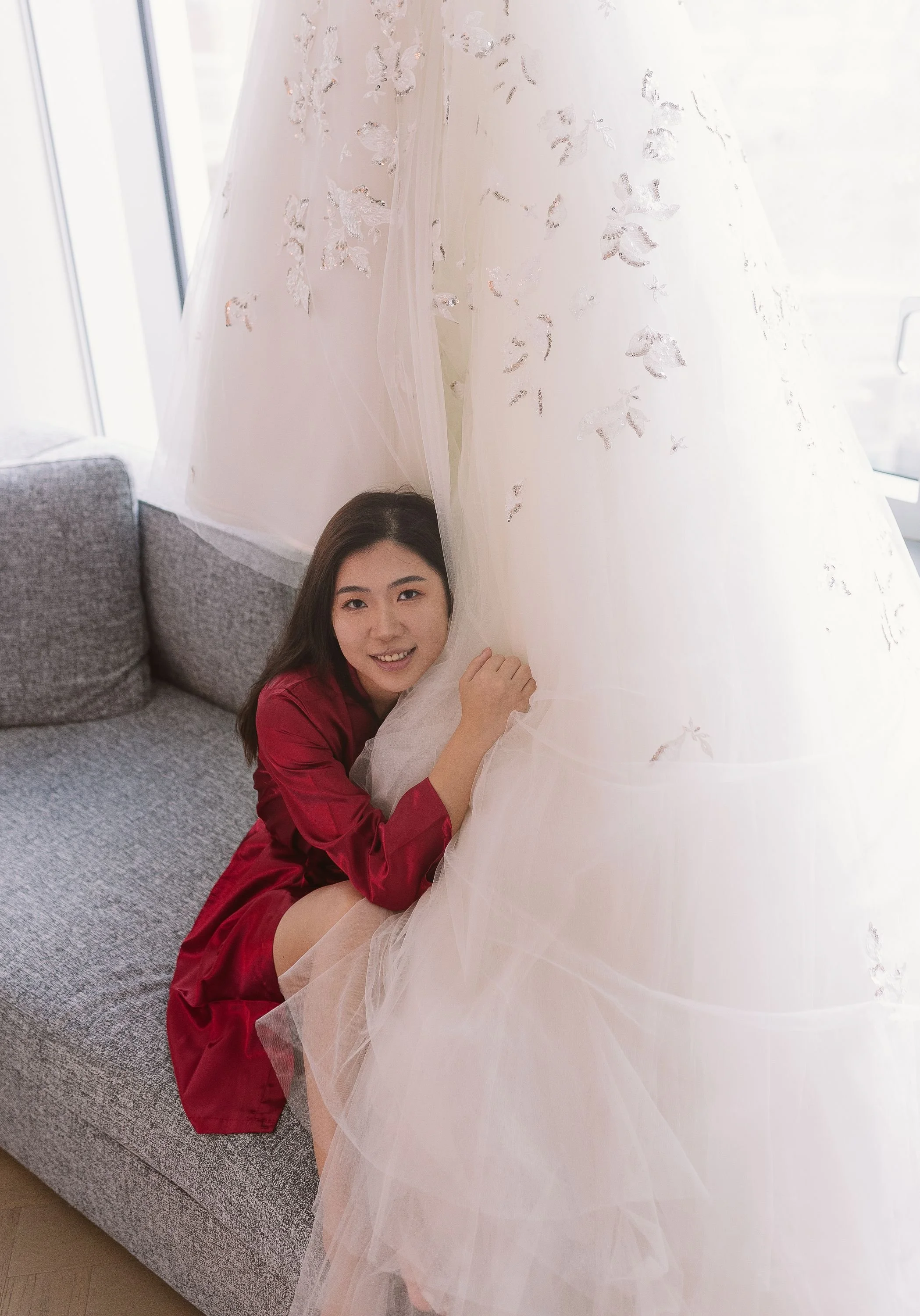 A woman with long dark hair, wearing a red satin dress, sitting on a gray couch, smiling and leaning against a large, white, floral-embellished wedding dress in a well-lit room with large windows.