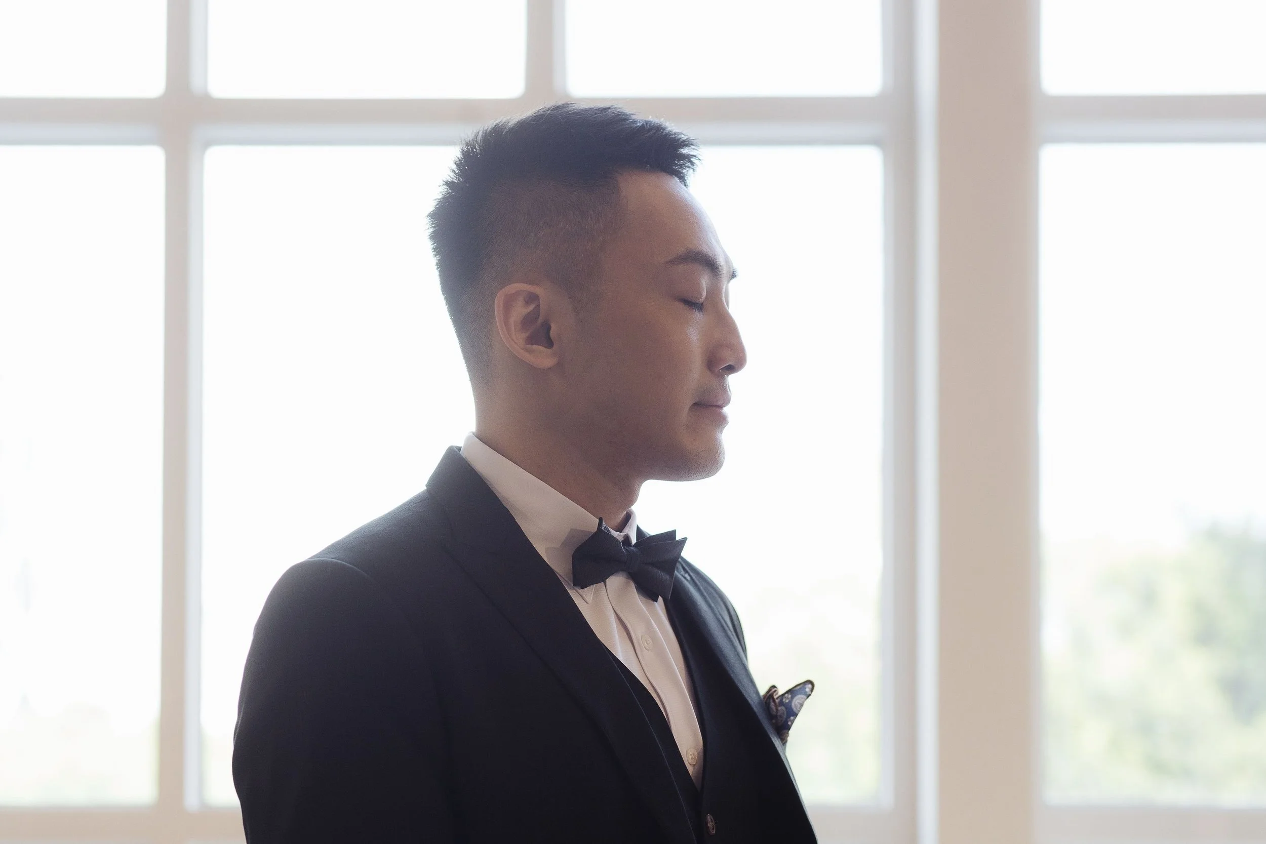 A man dressed in a black tuxedo with a bow tie, standing with closed eyes inside a room with large windows in the background.