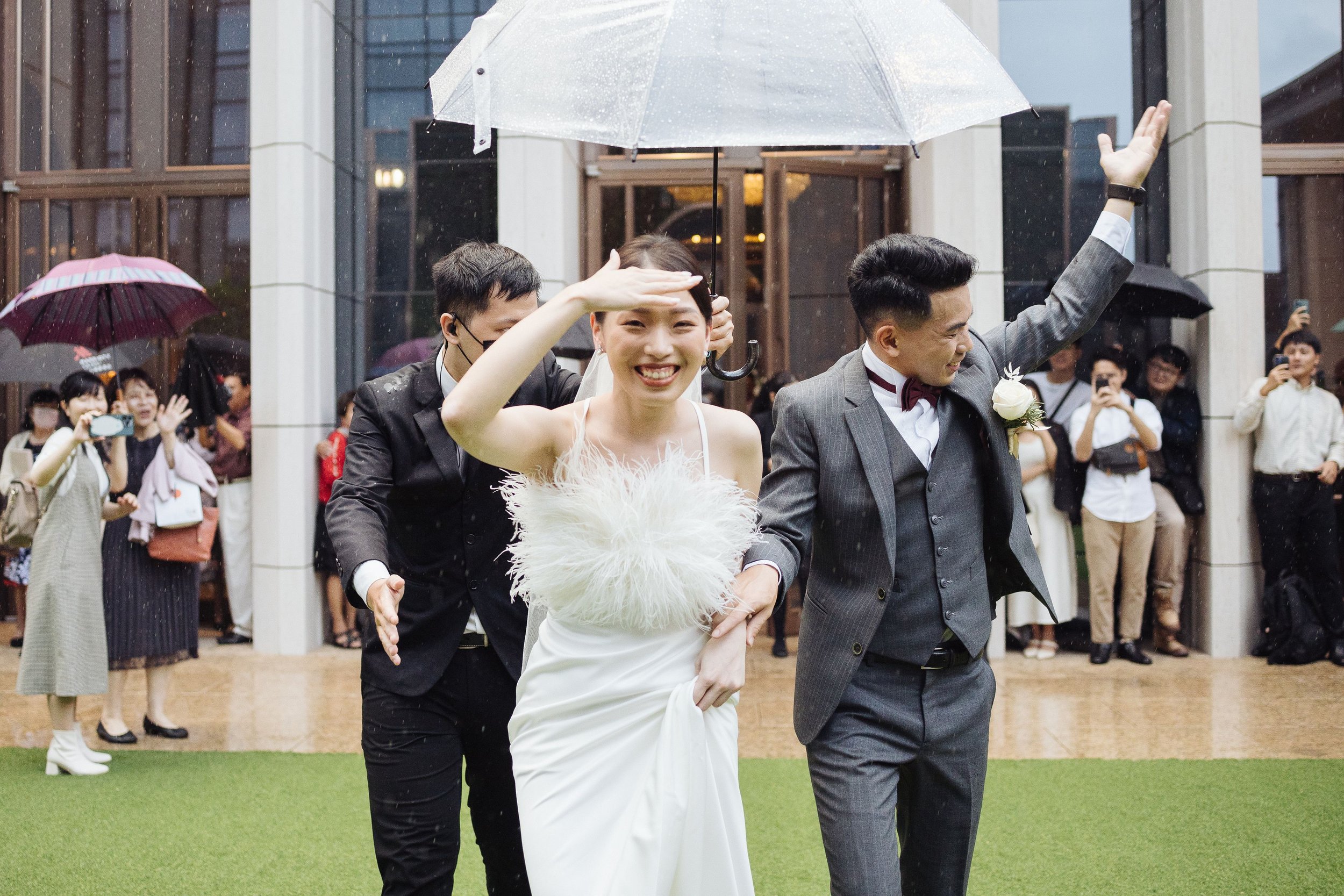 A bride and groom walking together outdoors during a wedding celebration in the rain, with friends and guests in the background taking photos and holding umbrellas.