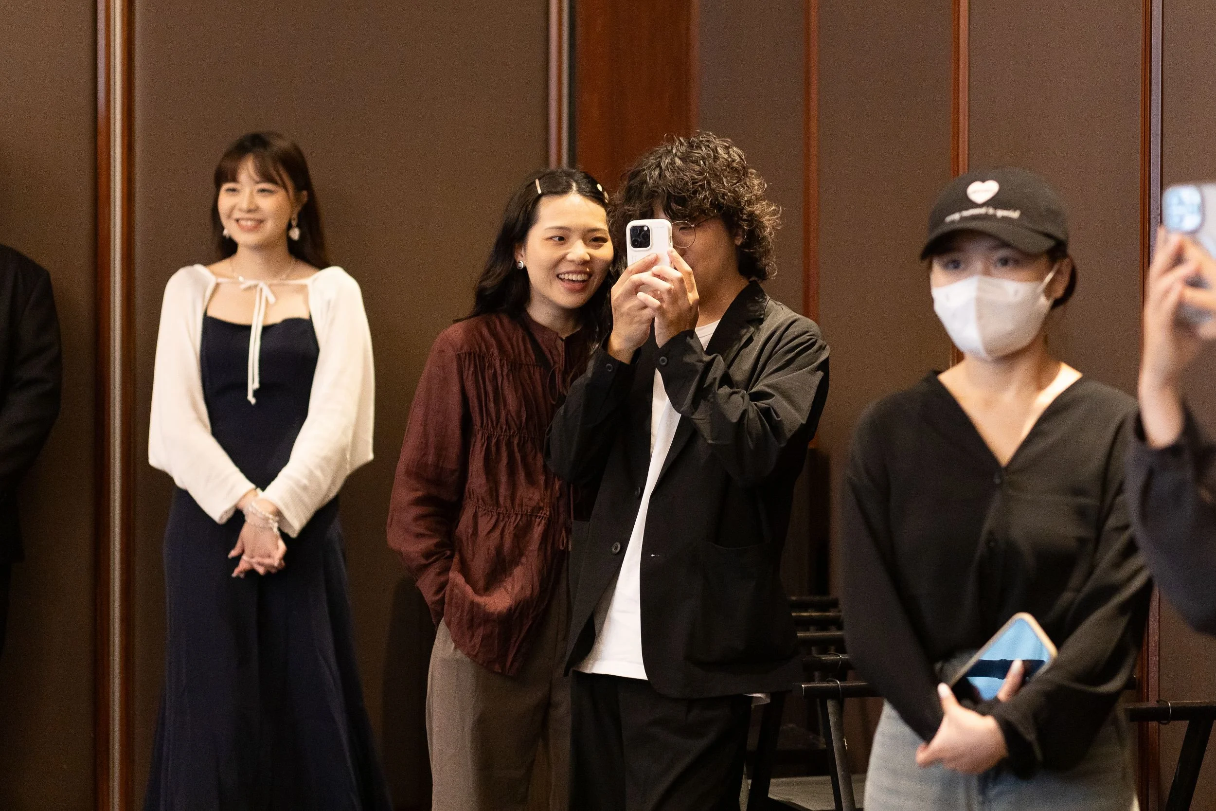 Four women are standing in an elevator or a room with wooden wall panels. One woman on the left is smiling, wearing a dark blue dress with a white cardigan. Two women in the middle are taking a selfie together, one with curly hair in a black jacket a