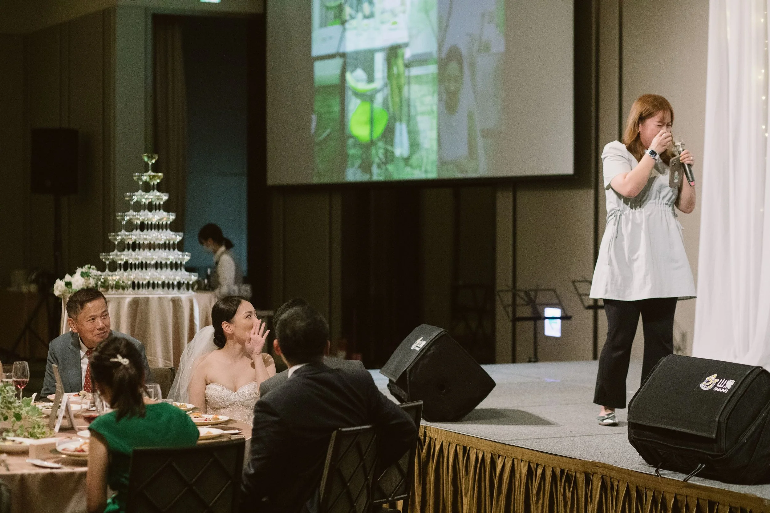 A woman with long hair is standing on a stage, holding a microphone and laughing. In front of her, seated at a table, are a bride in a white dress and a groom in a gray suit, along with other guests. There is a large decorative champagne tower, flora
