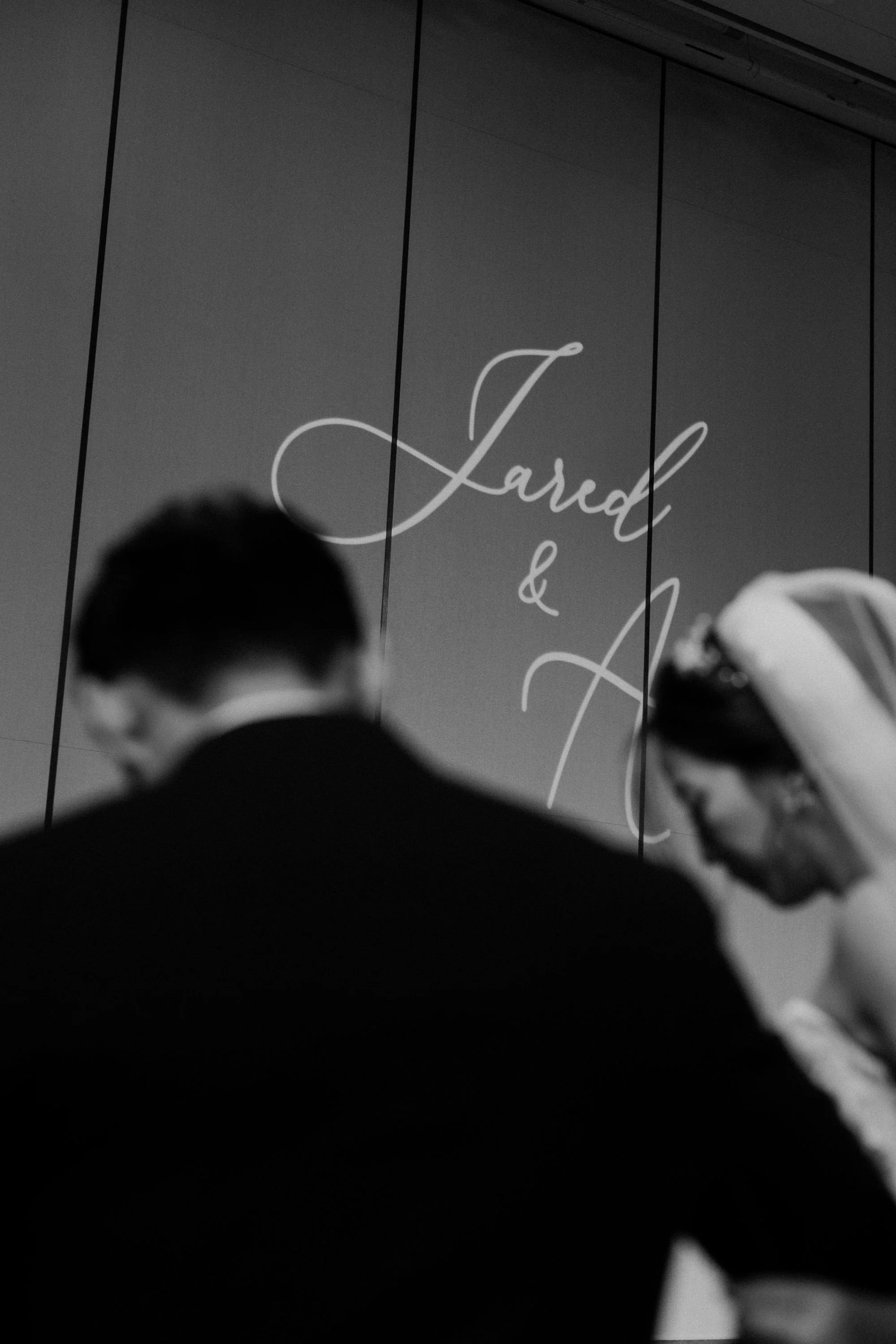 A black and white photo showing a projection on a wall with elegant cursive text that reads 'Lash &' and part of another word. In the foreground, three people are seen, two adults and one child, blurred and out of focus, attending an event or present