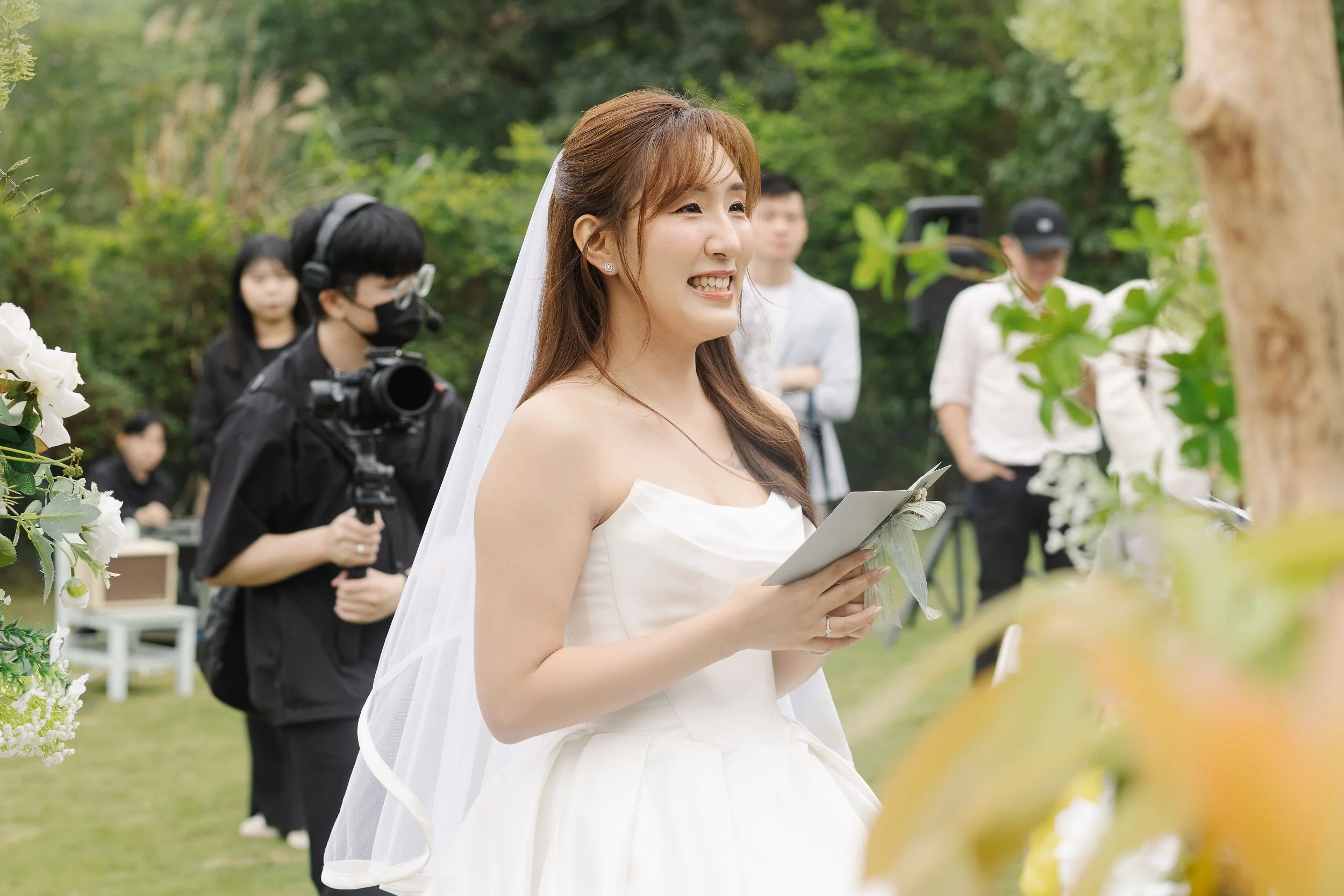 A smiling bride holding a bouquet and a card, standing outdoors during a wedding ceremony in a garden surrounded by greenery and flowers.