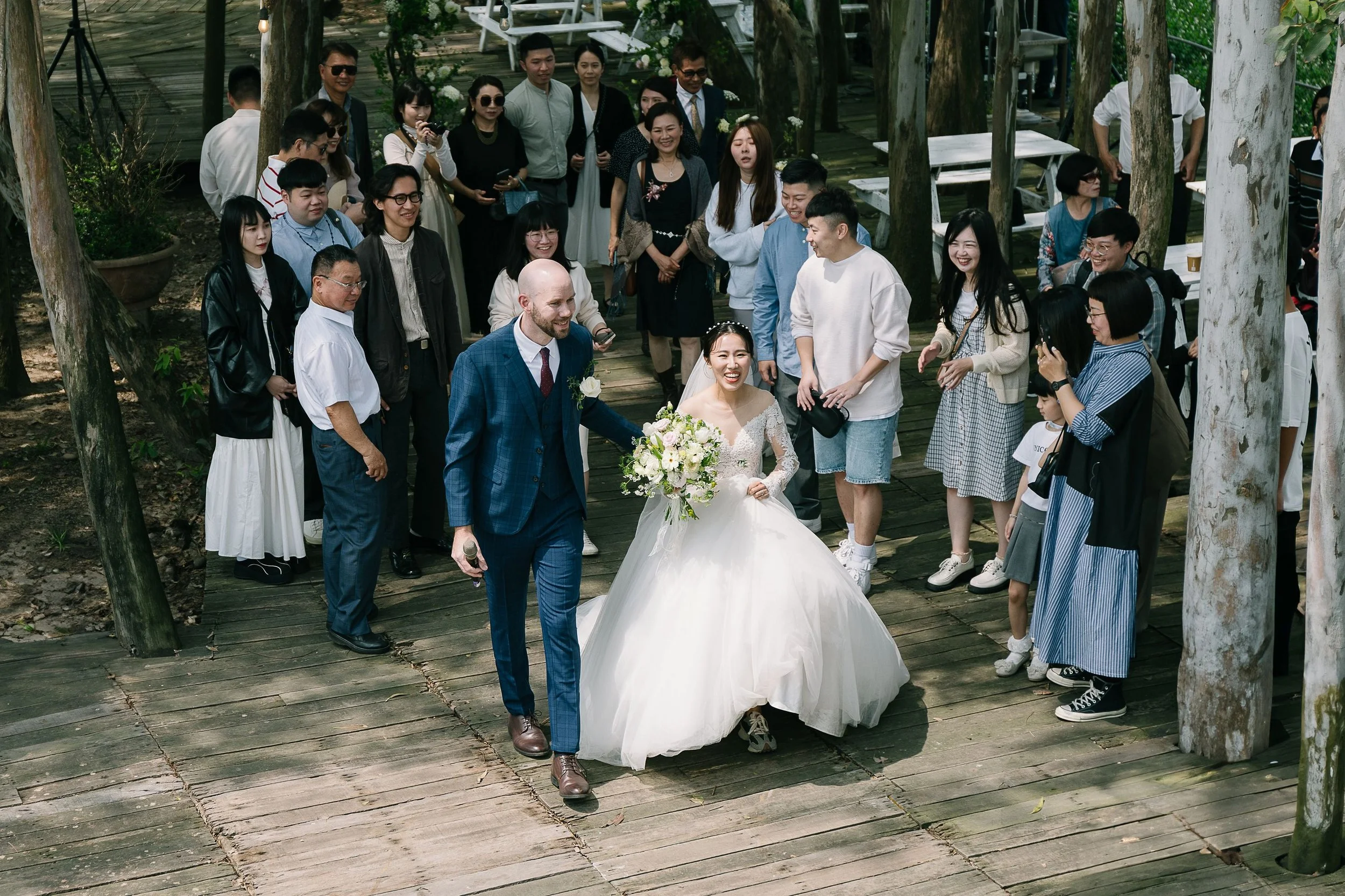 A wedding celebration outdoors on a wooden platform surrounded by trees, with the bride and groom walking together, smiling, while friends and family gather around, some taking photos and enjoying the moment.