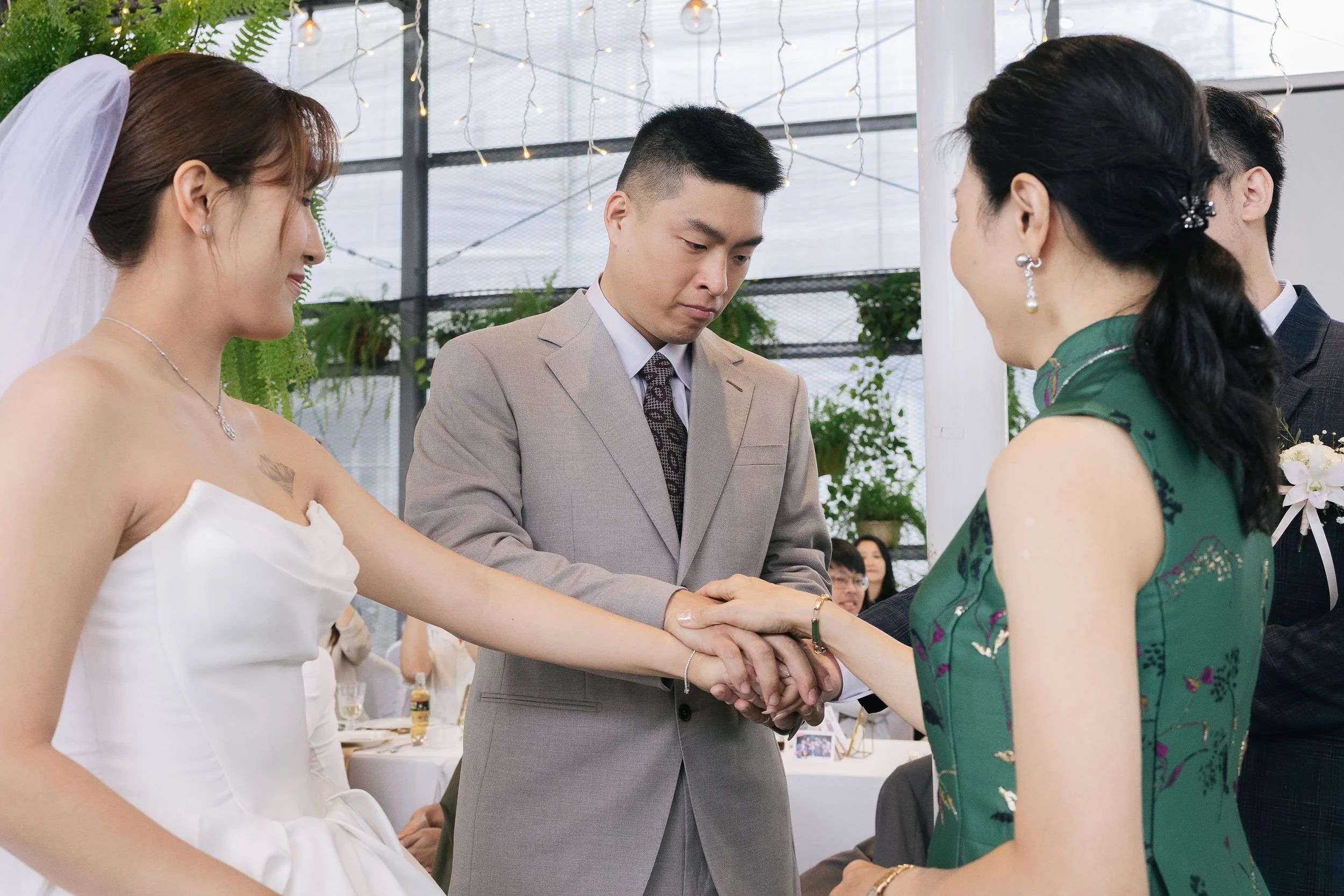 A wedding ceremony where a couple is exchanging vows, with a man in a tan suit and two women in dresses, one in white and the other in green. They are standing under decorative string lights, holding hands, with a floral bouquet visible in the backgr