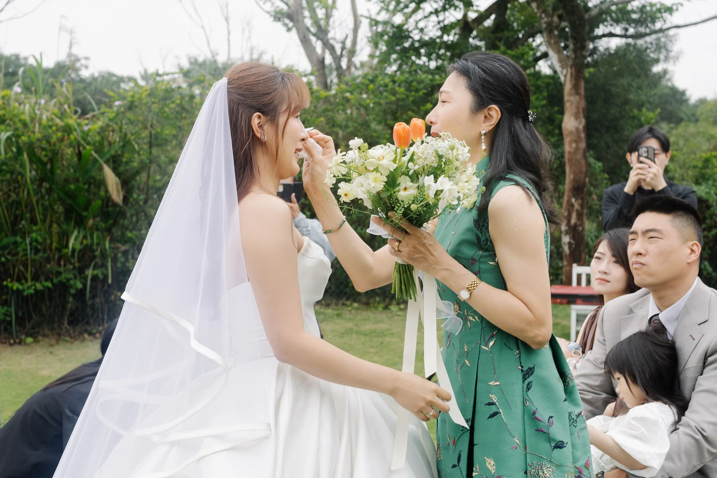 A bride in a wedding dress and veil is being handed a bouquet of flowers by a woman wearing a green dress at an outdoor wedding ceremony.